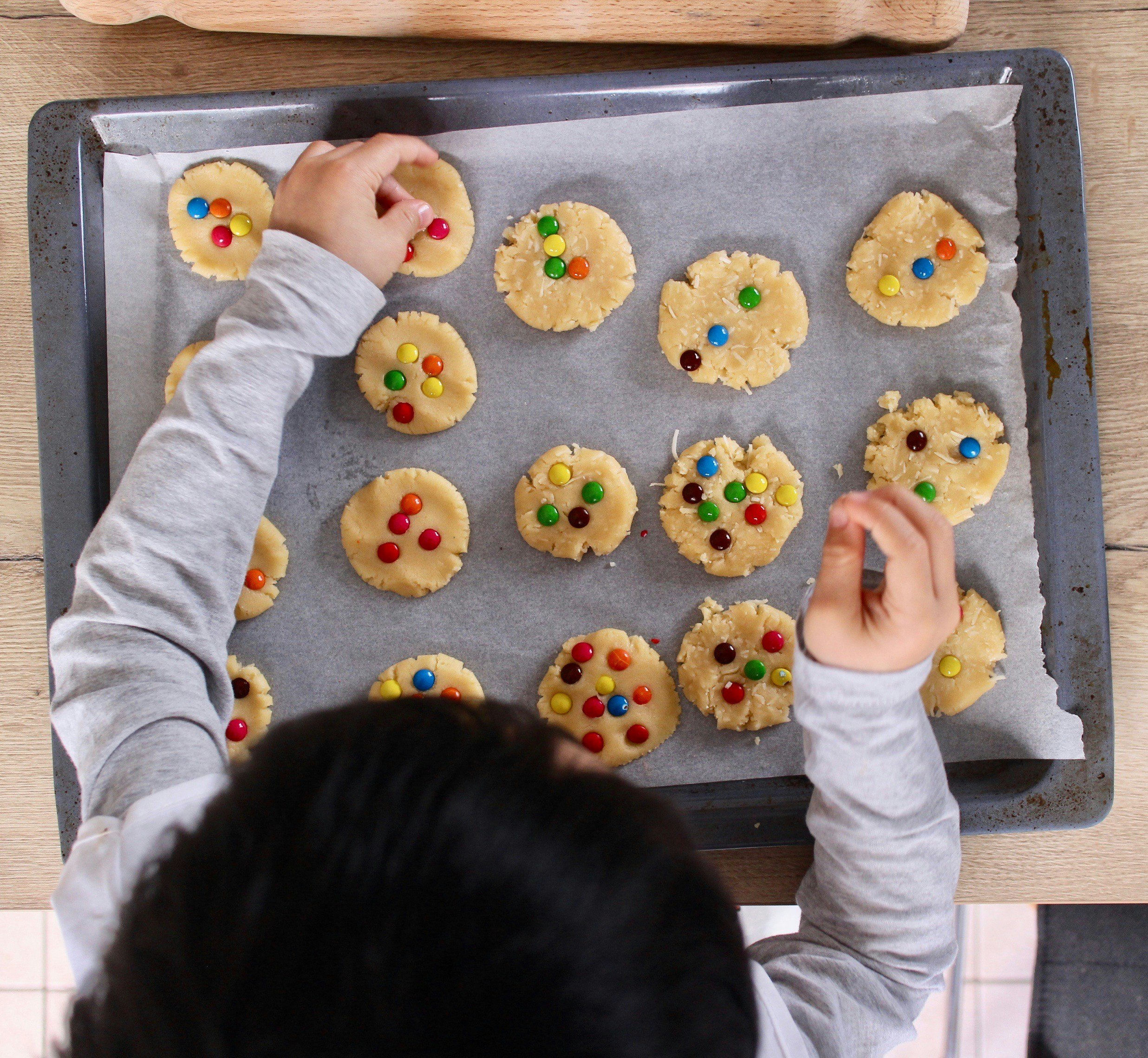 Child arranging cookie dough with colorful candies on a baking sheet covered with parchment paper.