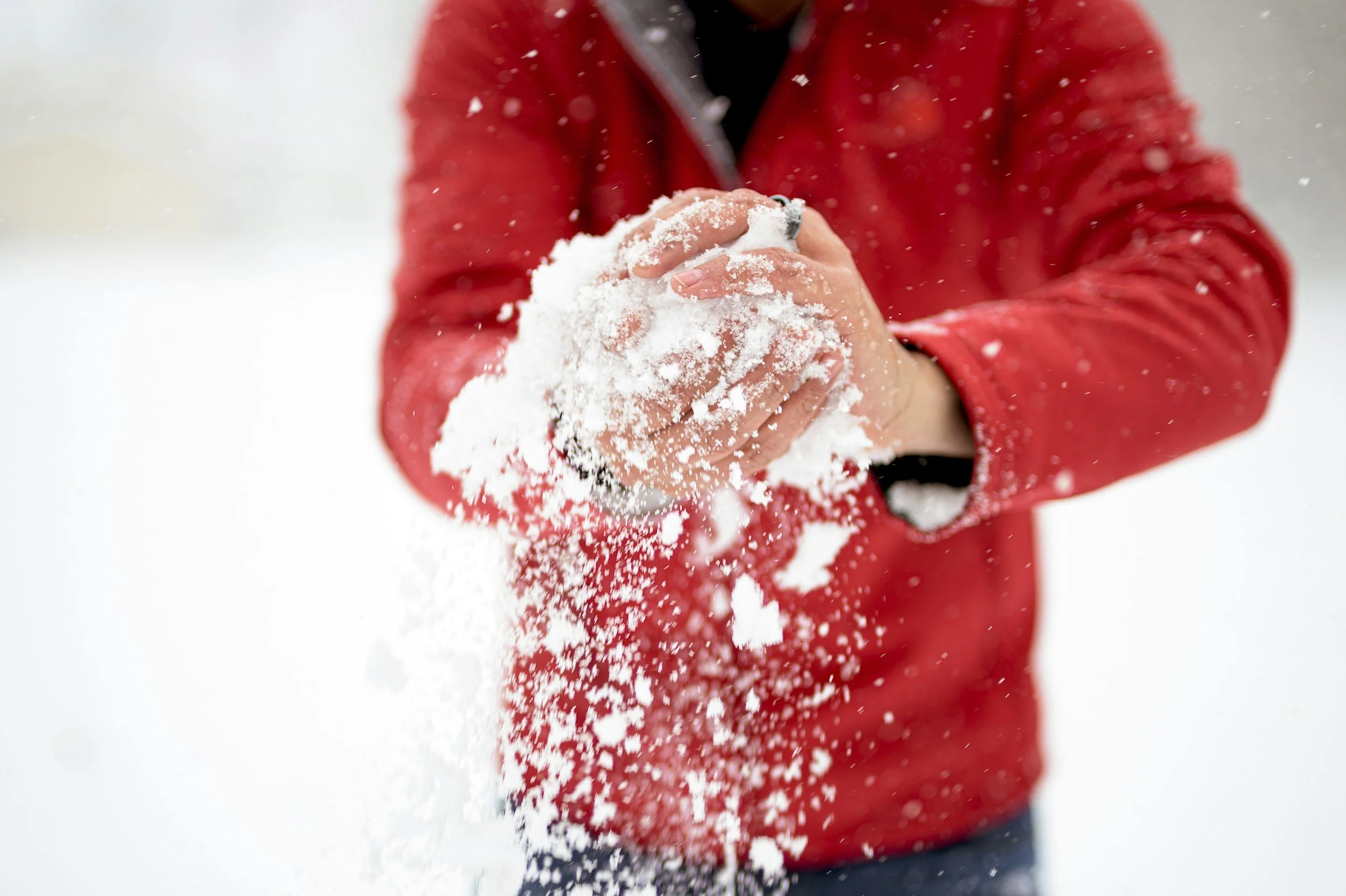 A person wearing a red jacket is holding snow in their hands, throwing snow into the air outside.