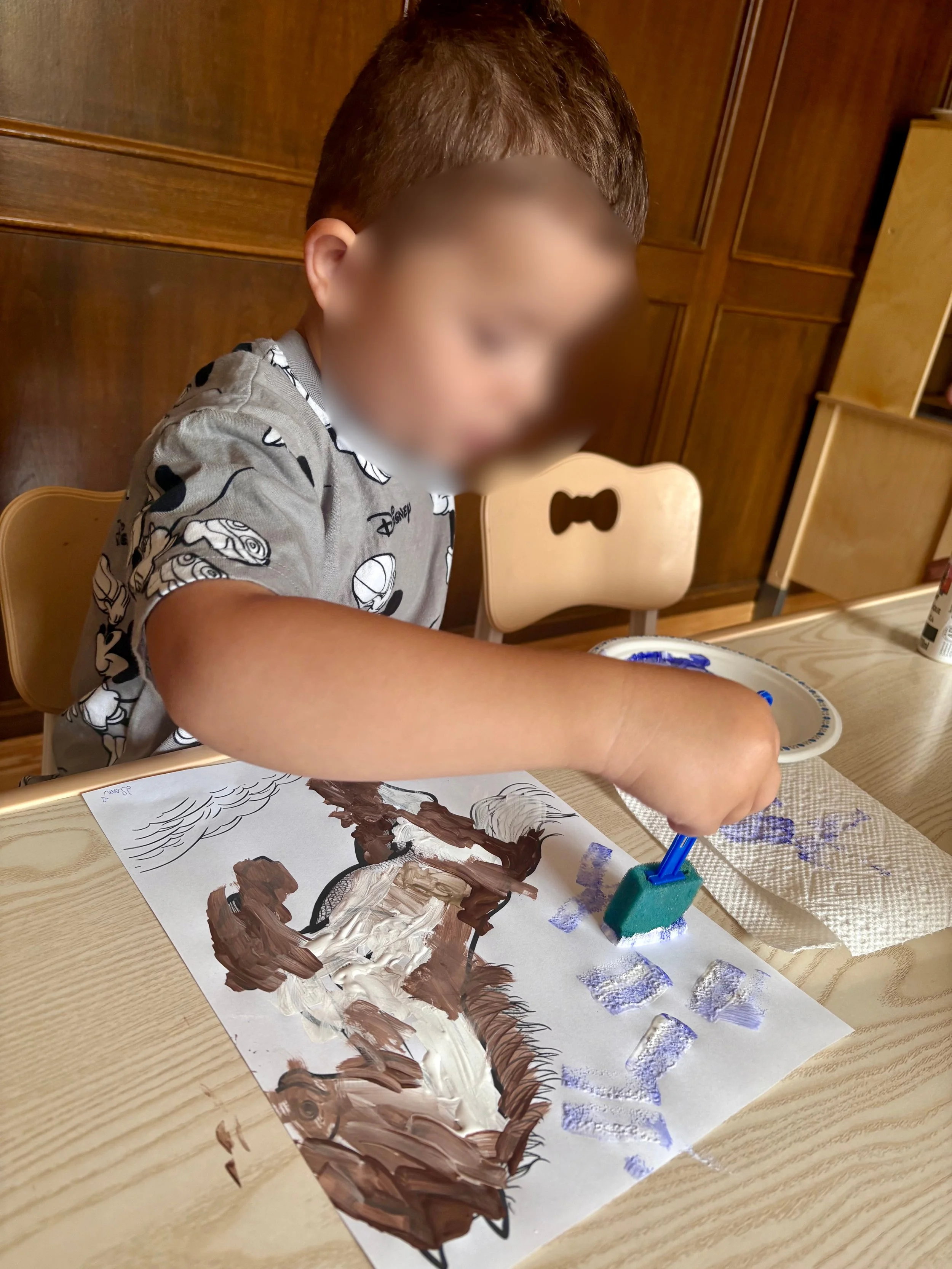 A young child painting a picture of a dog with thick brown and white strokes on a sheet of paper.