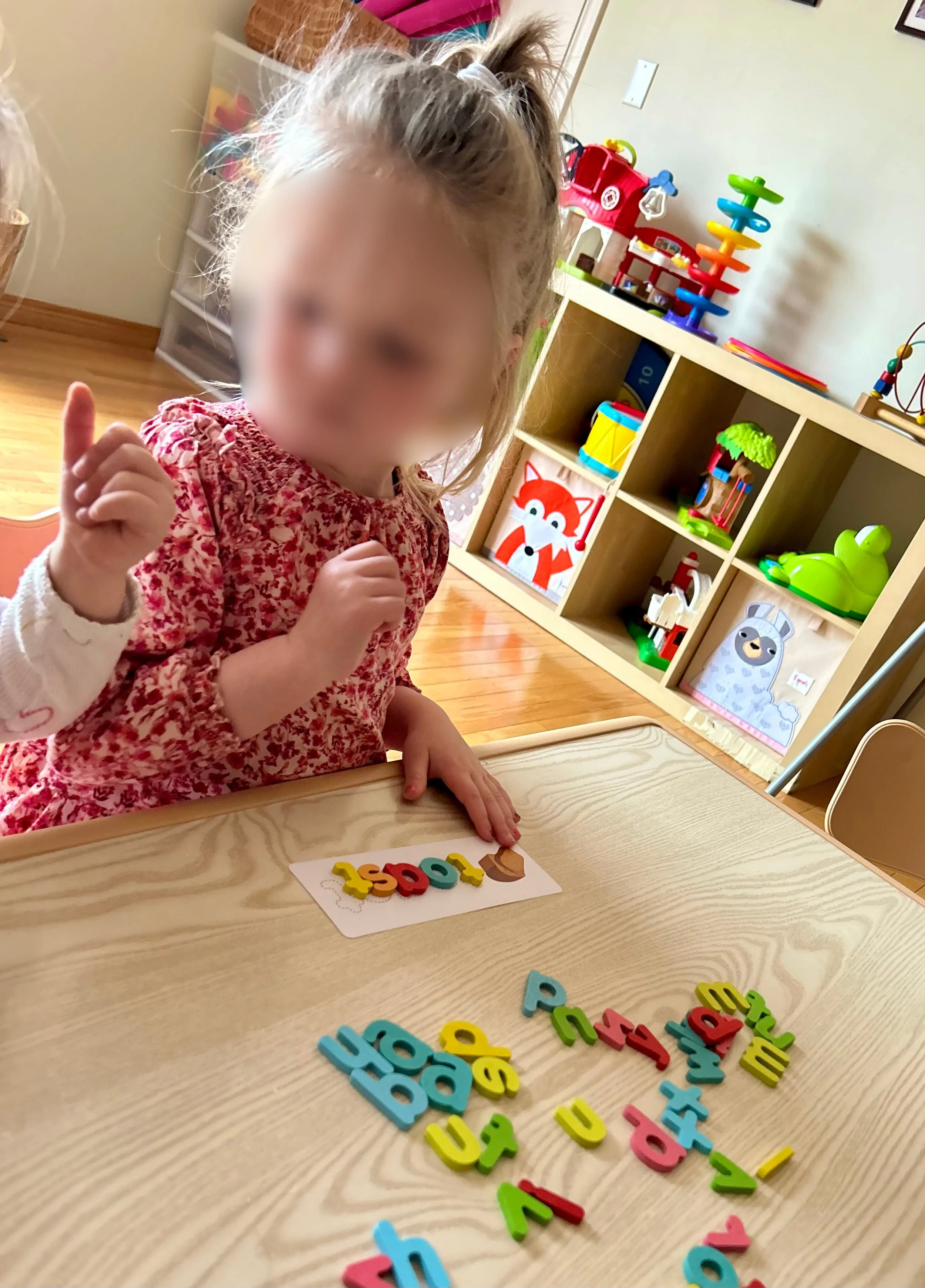 A young girl with blonde hair tied in a ponytail, sitting at a wooden table, working on a learning activity with colorful magnetic or foam letters. The background shows a toy shelf with various toys, including a princess castle, a fox picture, and ot