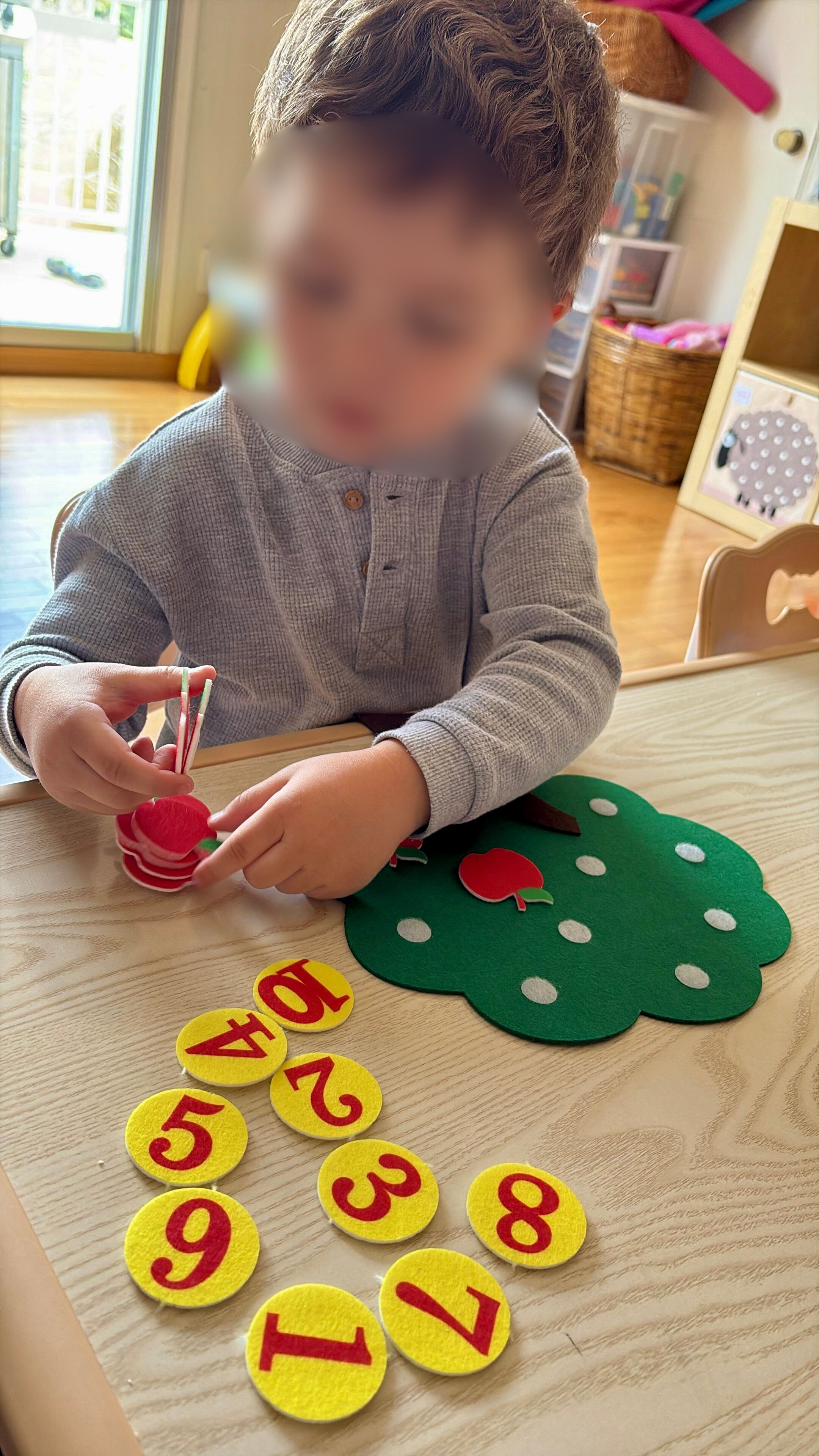 A young child with blurred face playing with felt apple and leaf shapes, organizing yellow felt circles with numbers in red. The circles display numbers 1 to 10, along with a circle labeled '10+'. The child is seated at a wooden table inside a cozy r