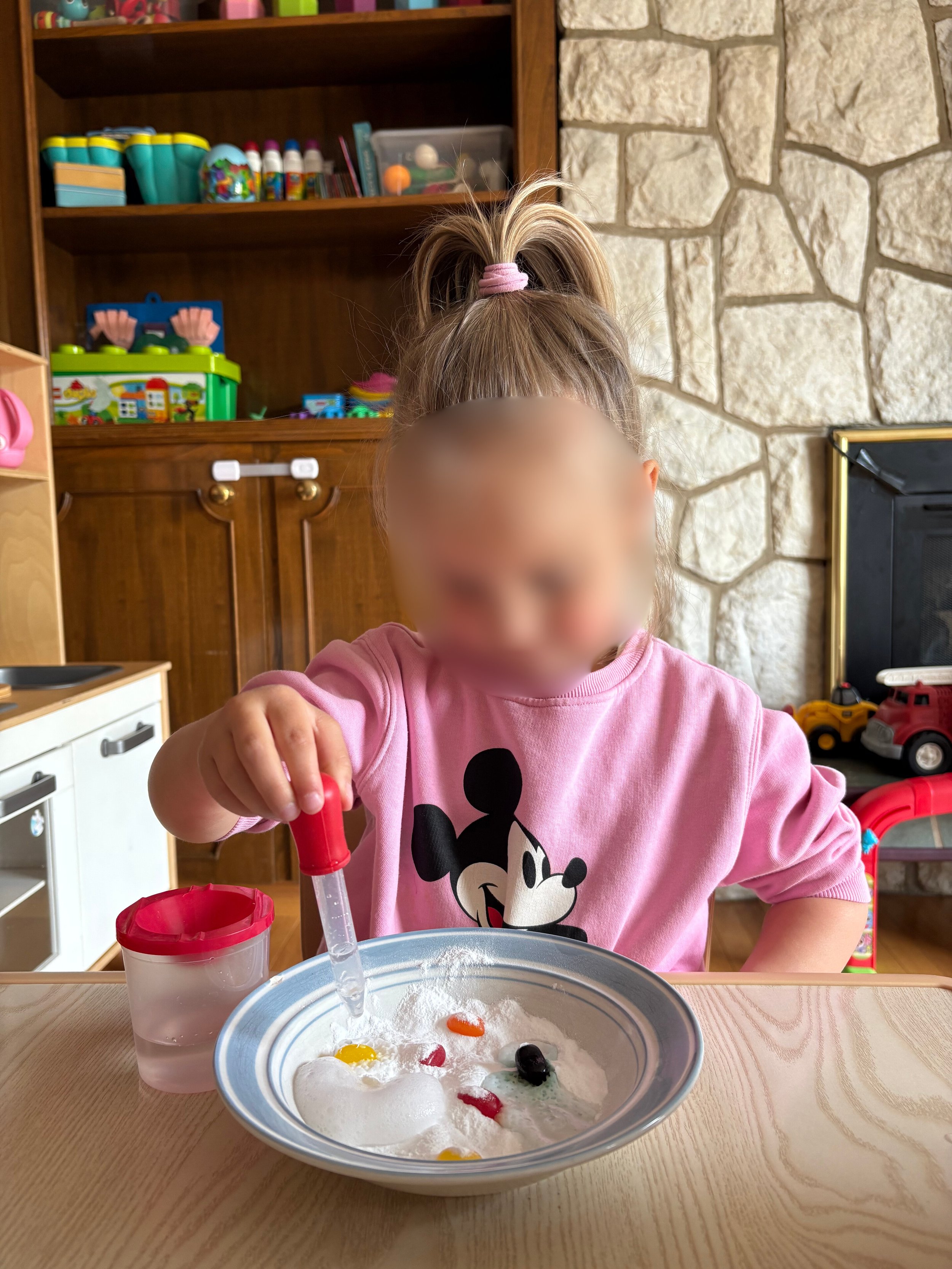 A young girl with a ponytail tied with a pink hair tie, wearing a pink Mickey Mouse t-shirt, is engaging in a science experiment with a bowl of baking soda and food coloring, using a dropper. There is a clear cup with water beside her on a wooden tab