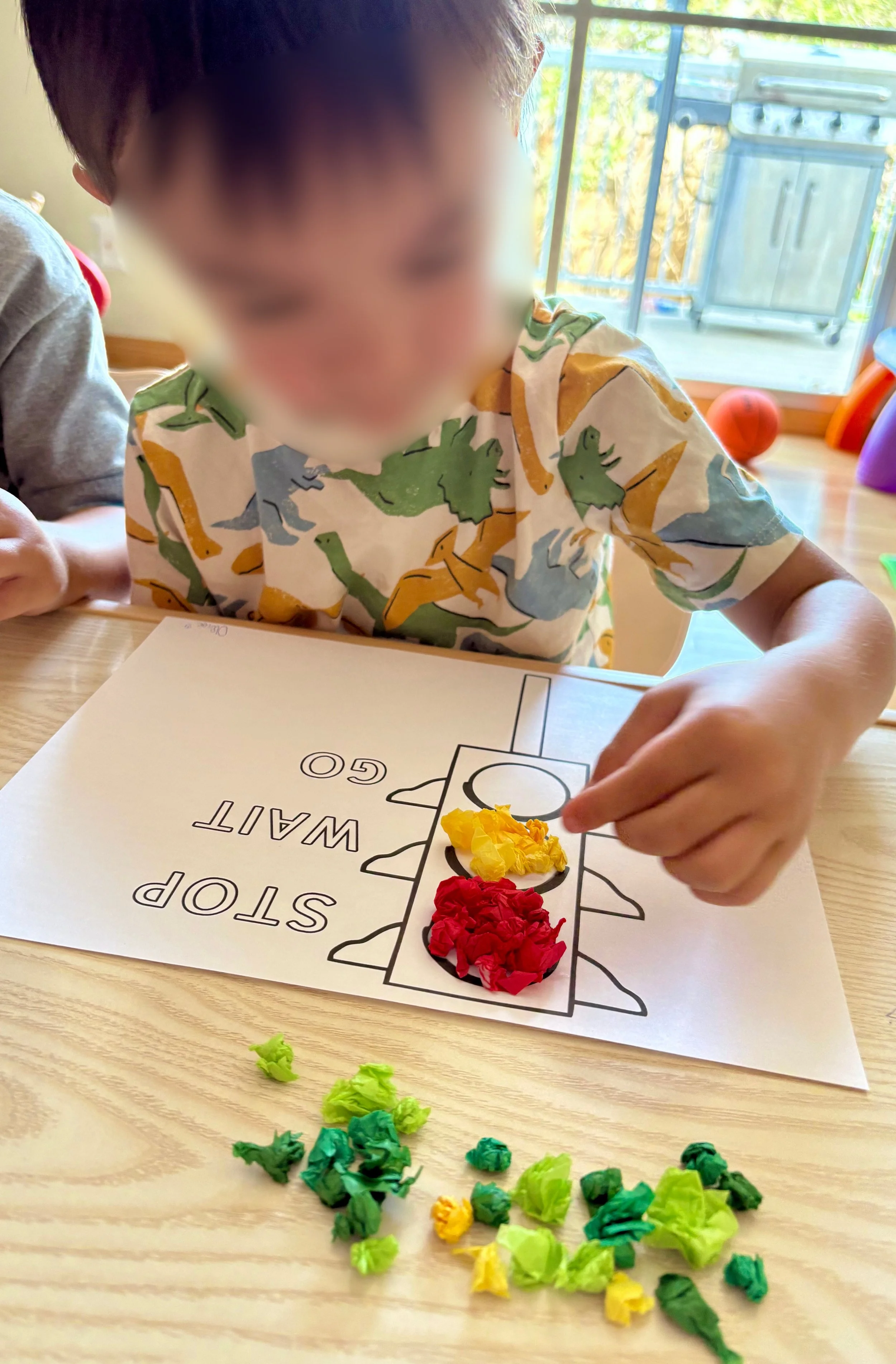 A child wearing a dinosaur-print shirt is placing red crumpled paper on a guided traffic light activity worksheet, with yellow and green crumpled paper on the table in front, inside a bright room with a sliding glass door leading outside.
