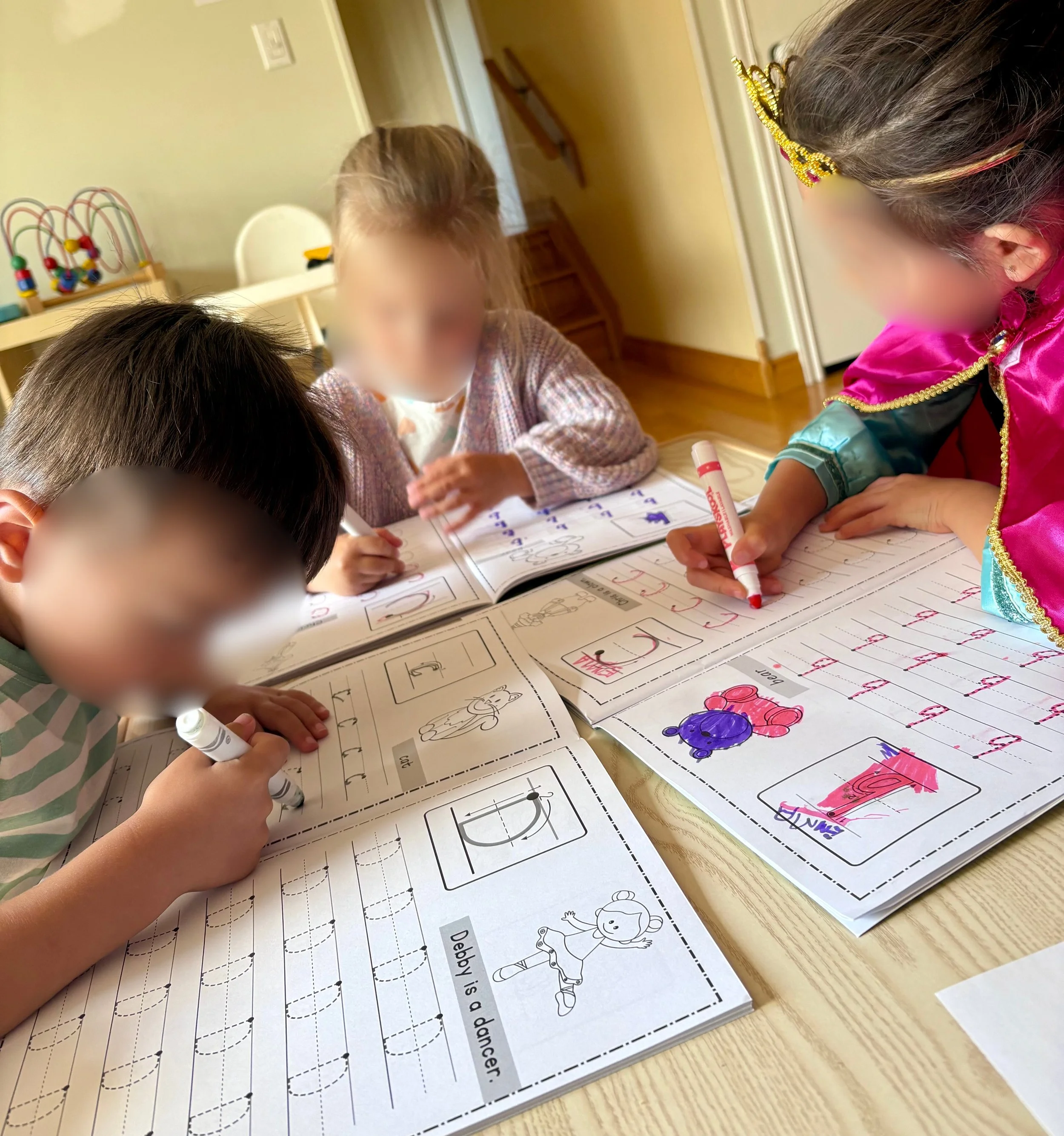 Four children at a table practicing writing in workbooks with ABC letters, markers, and drawings on a wooden table.