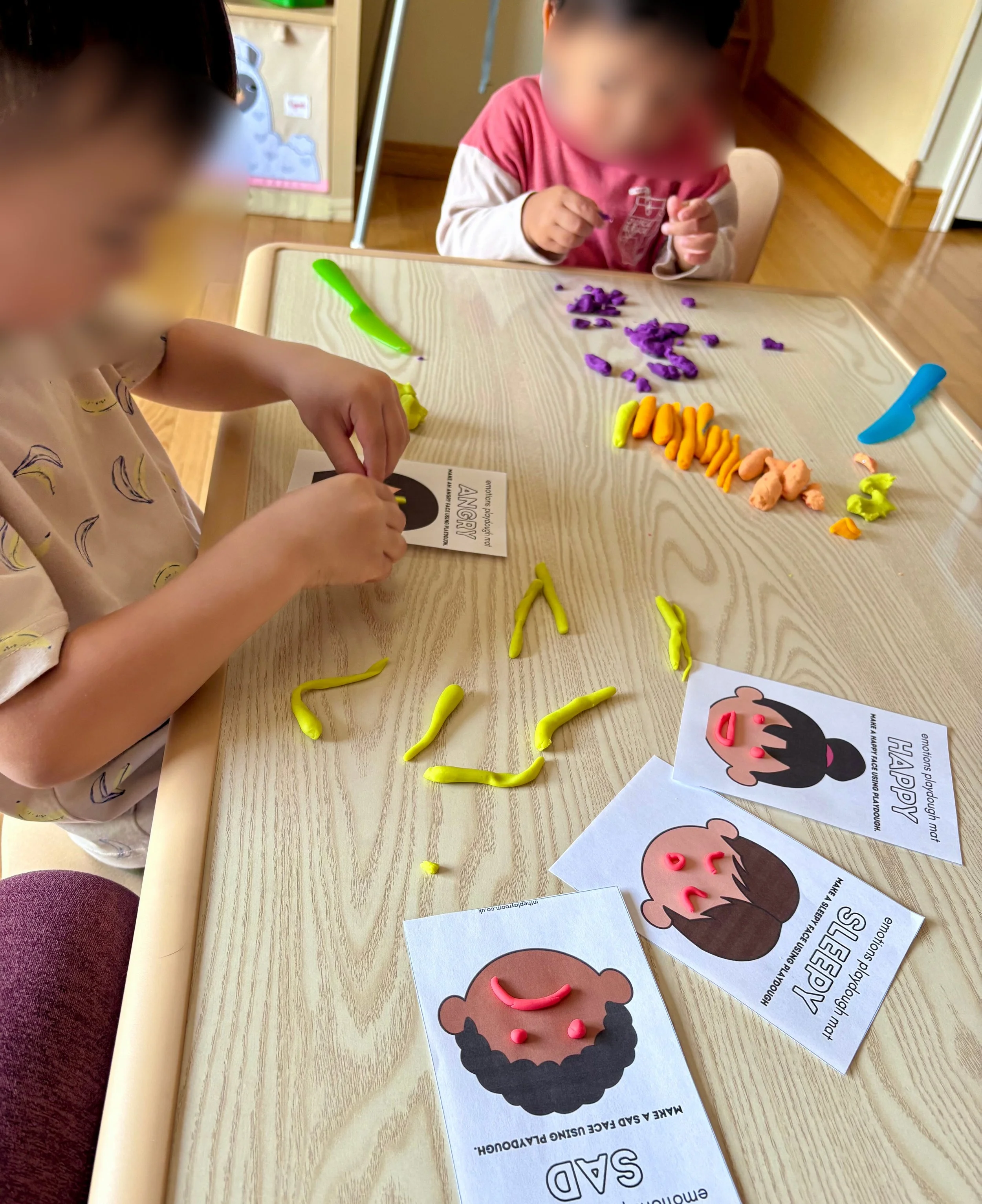 Children engaged in sensory play with colorful modeling clay, creating faces on printed templates with paper eyes, mouth, and cheeks, on a wooden table.