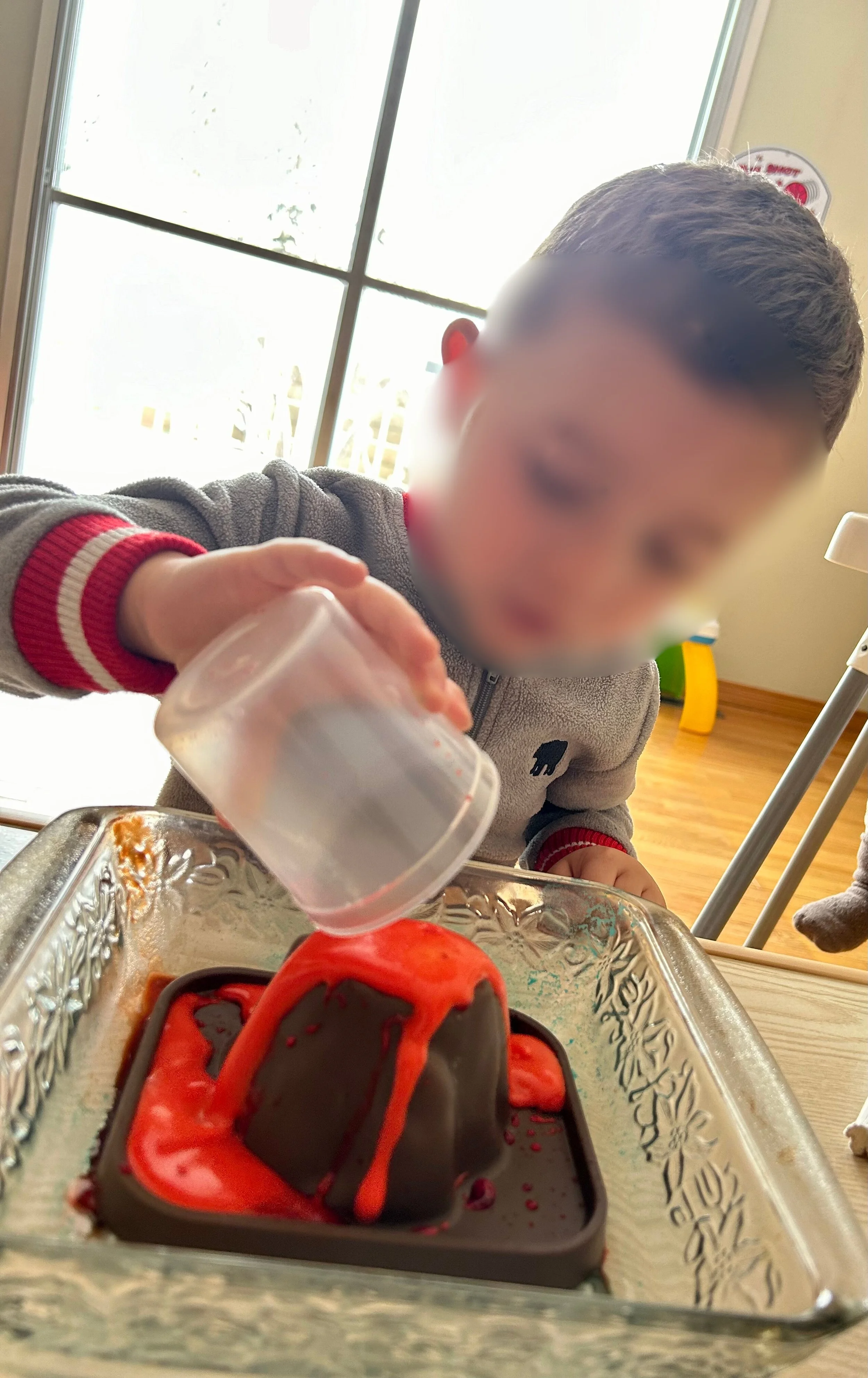 A young child with blurred face decorating a chocolate lava cake with red icing, using a small clear cup, in a kitchen or dining area with a window in the background.