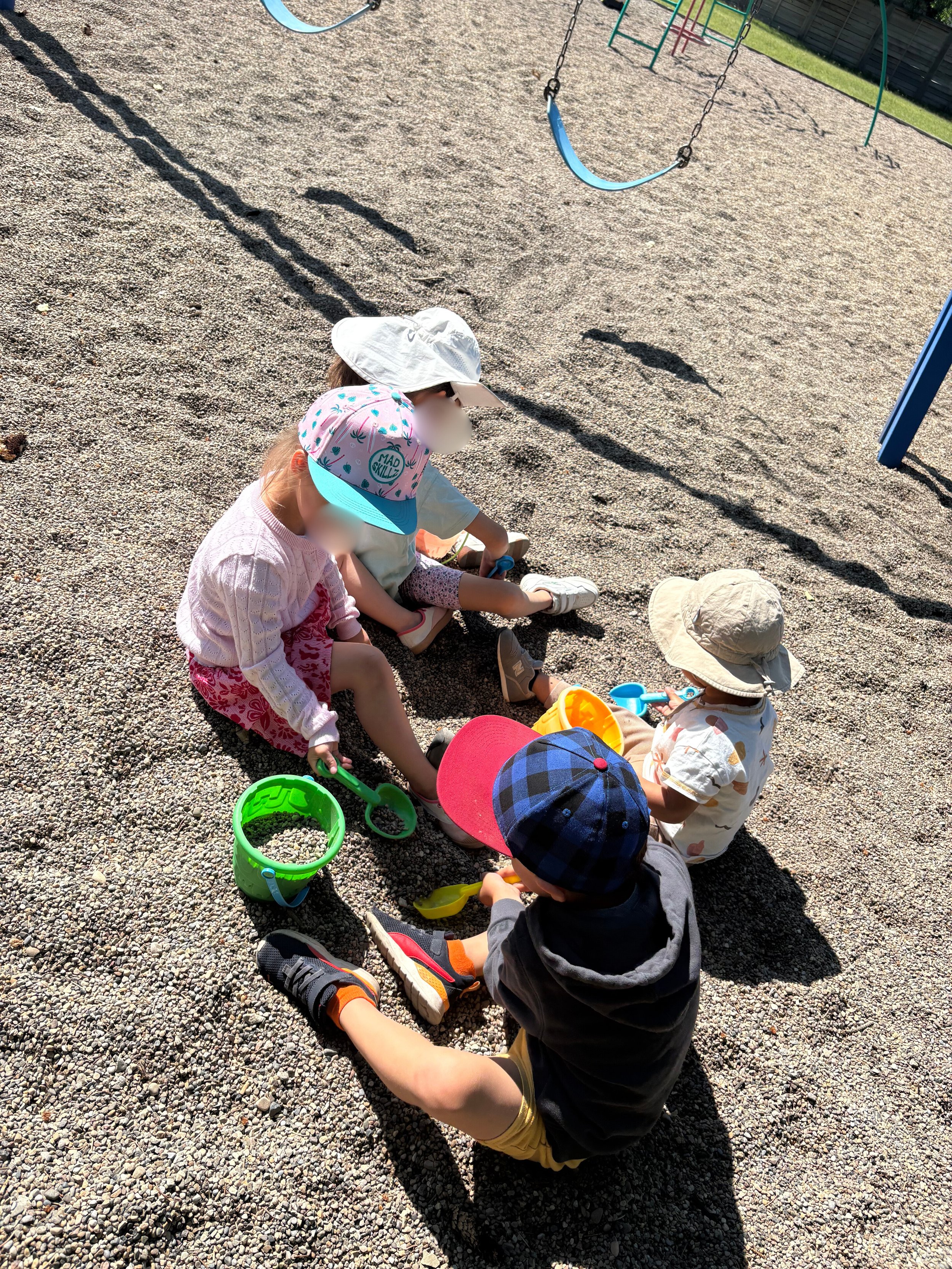 Four children sitting on gravel ground in a playground, playing with buckets and toy shovels. They are wearing hats and casual clothes, with a swing set visible in the background.