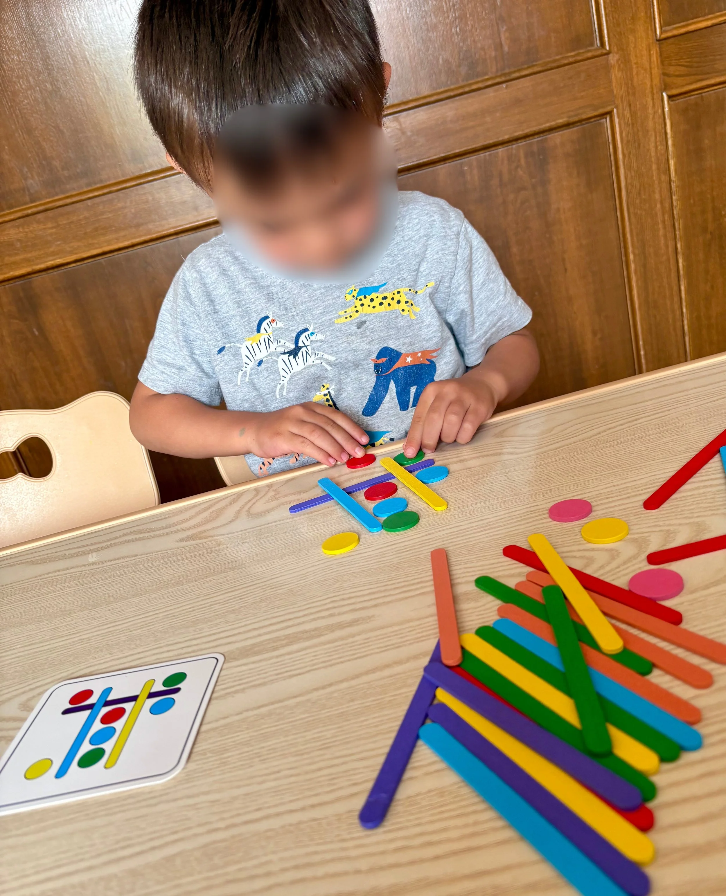 A young child playing with colorful sticks and tokens at a wooden table with a card game on the surface.