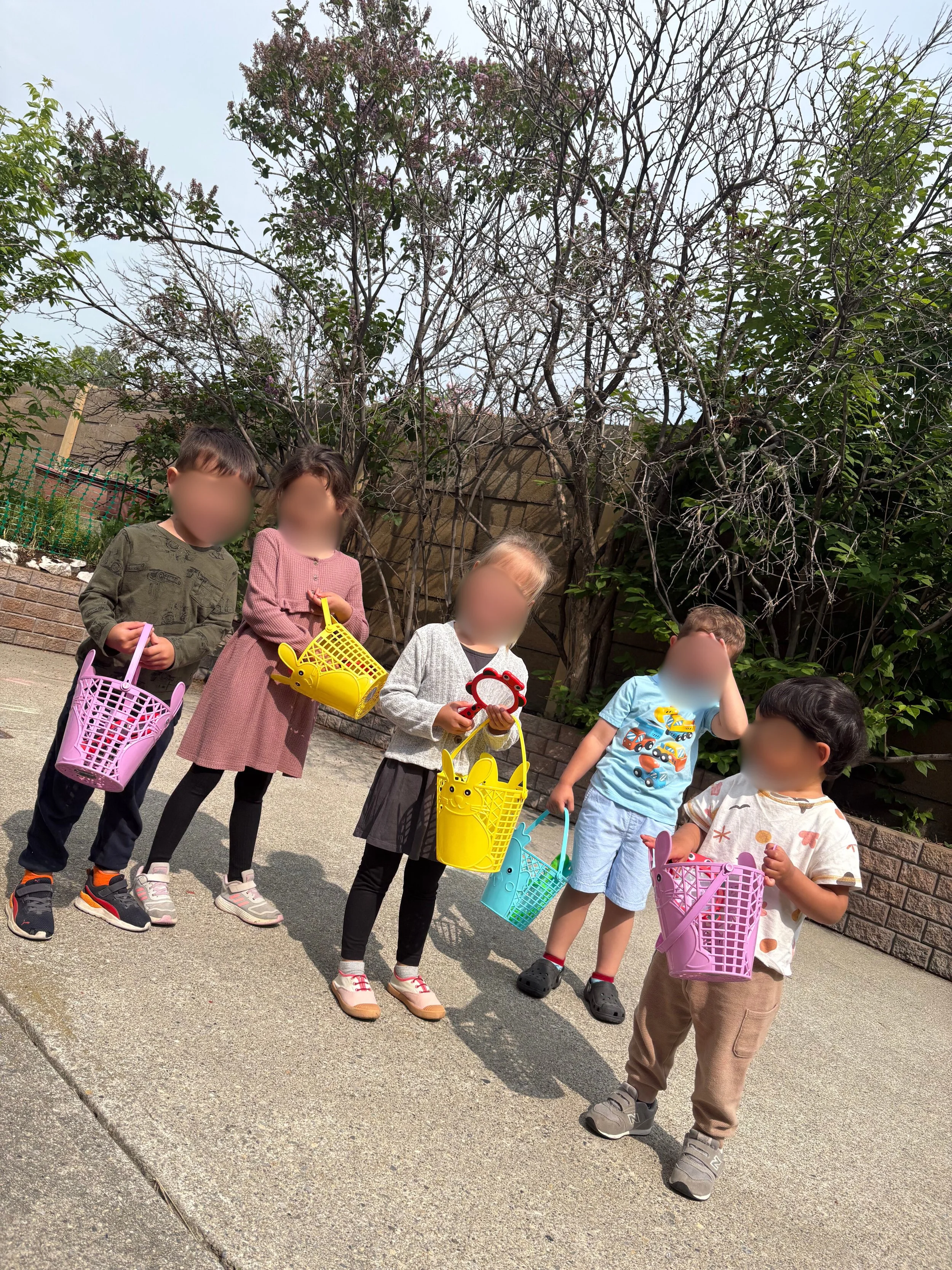 Six children standing outdoors holding colorful Easter baskets, with trees and a brick wall in the background.