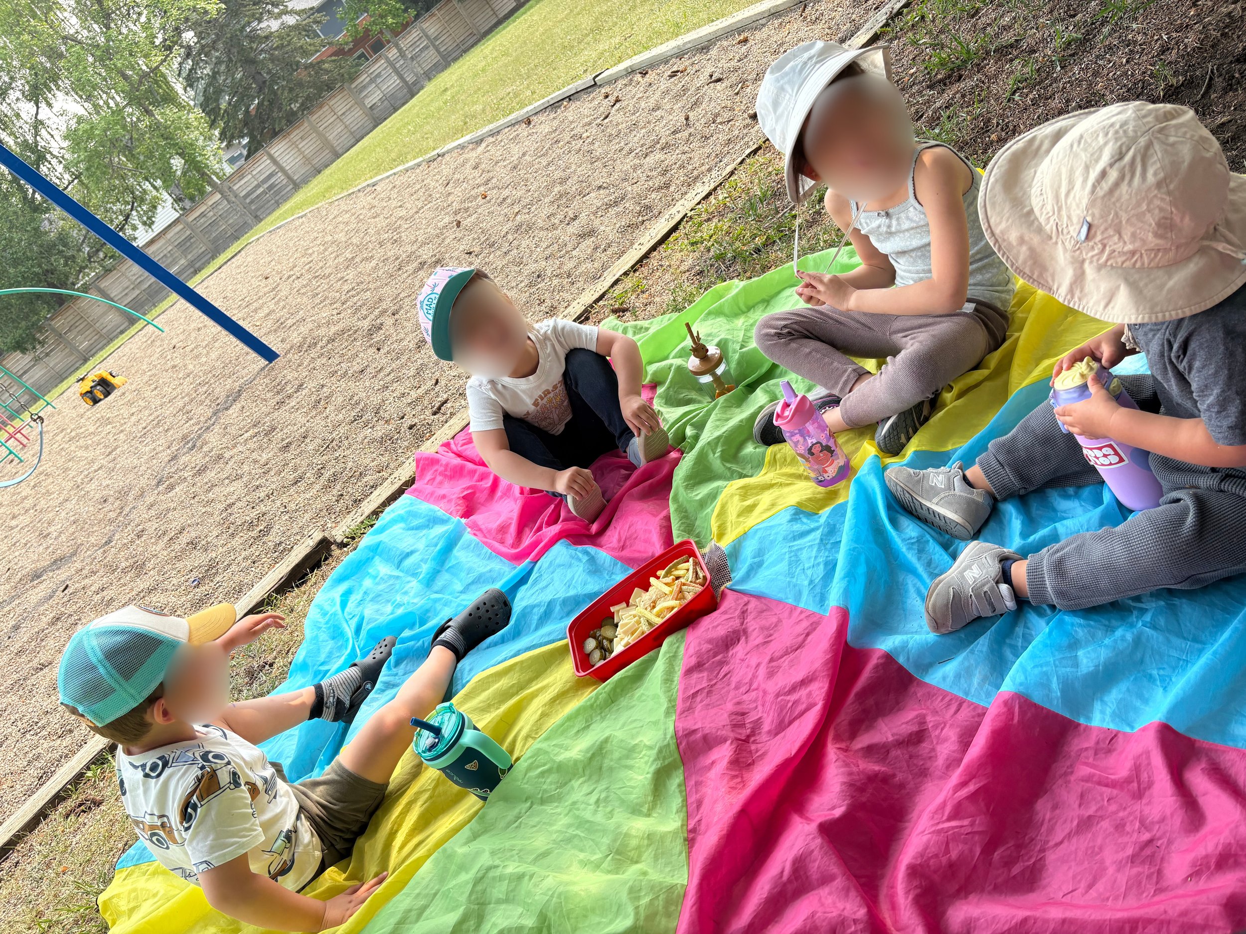 Four children sitting on a colorful blanket having snacks in a park.