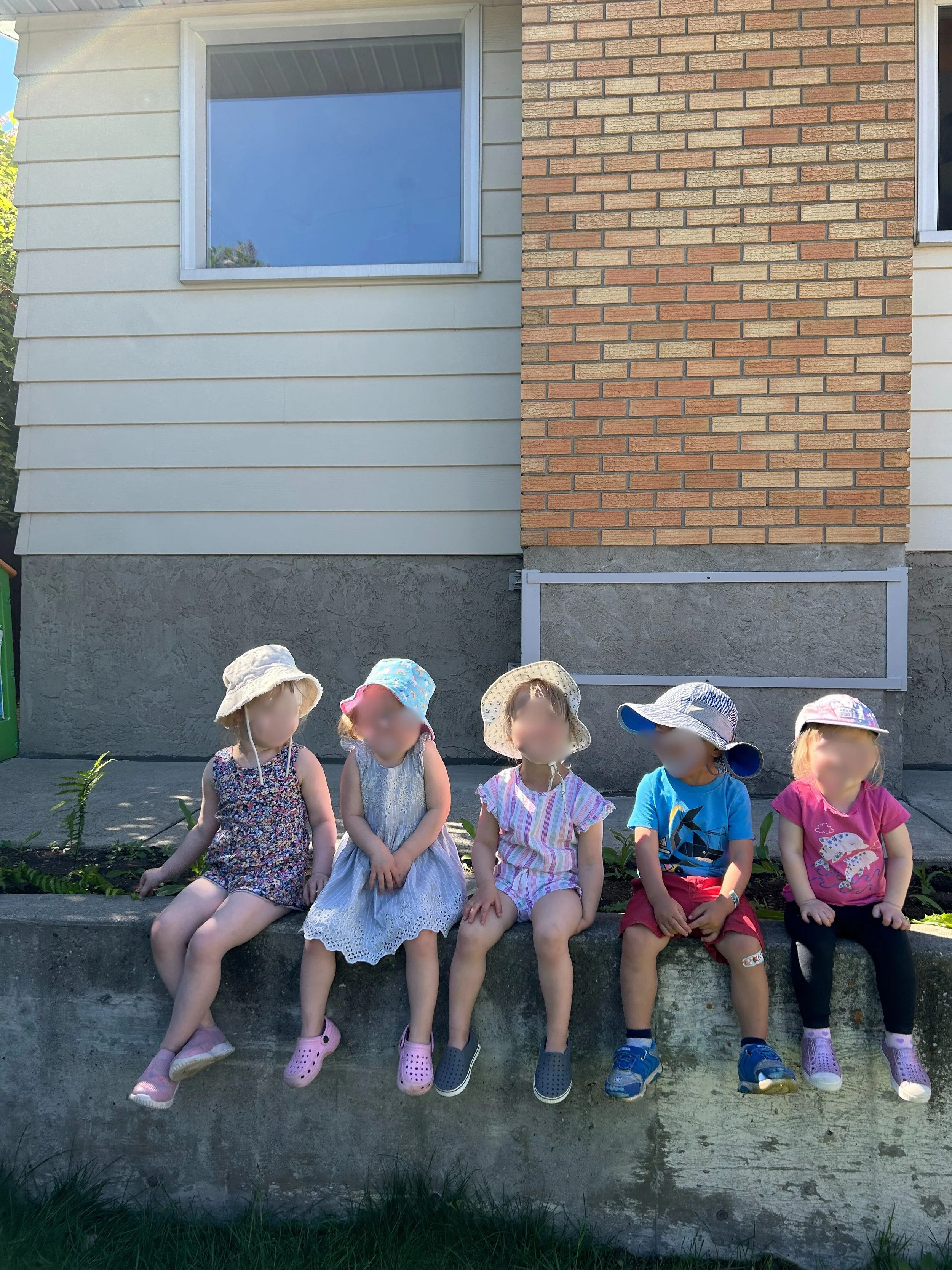 Five young children sitting on a concrete ledge outside a house, wearing summer clothes and hats.