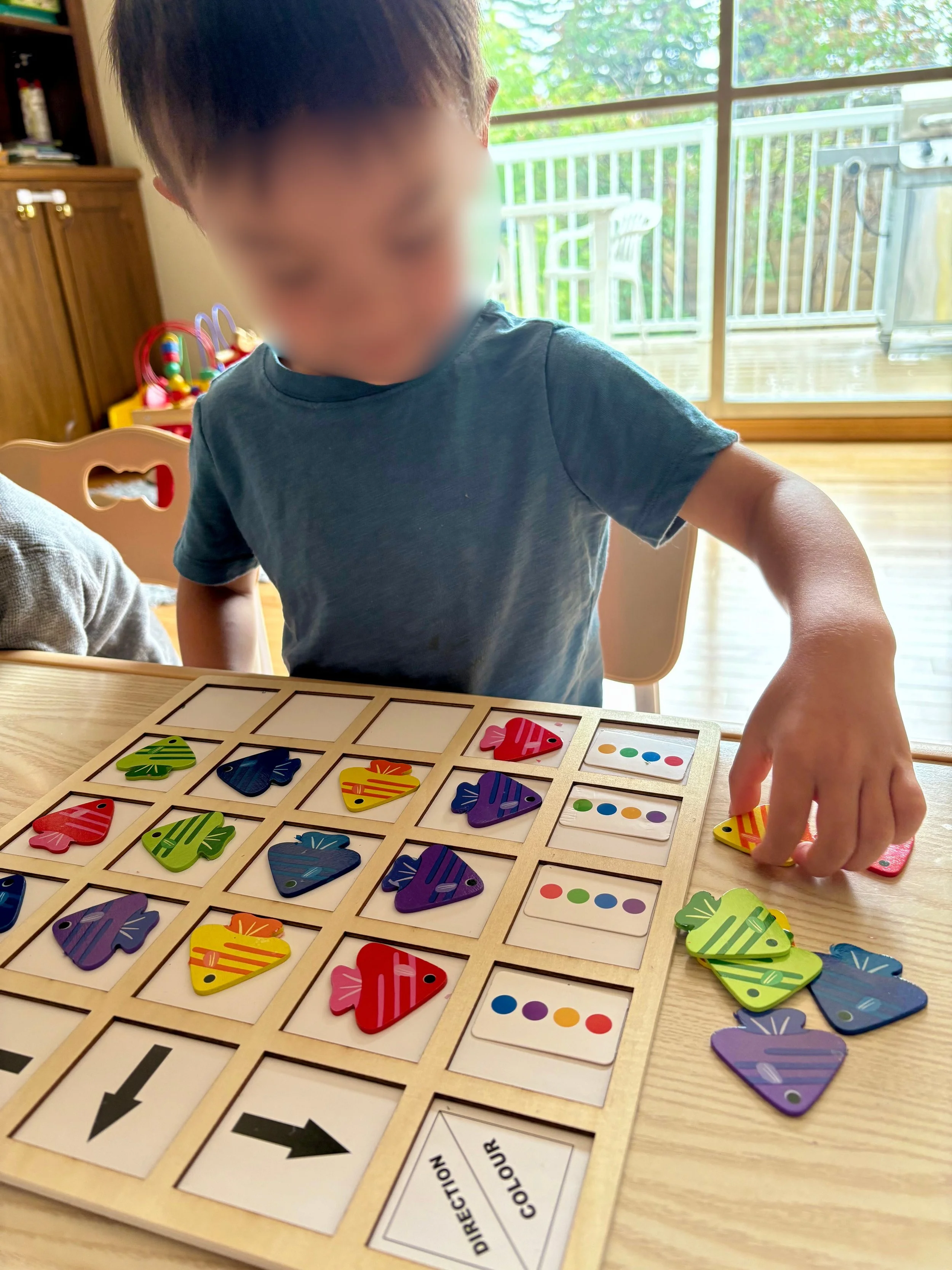 Child playing a board game with colorful fish-shaped tiles and direction cards on a wooden table in a room with a sliding glass door and outdoor deck.