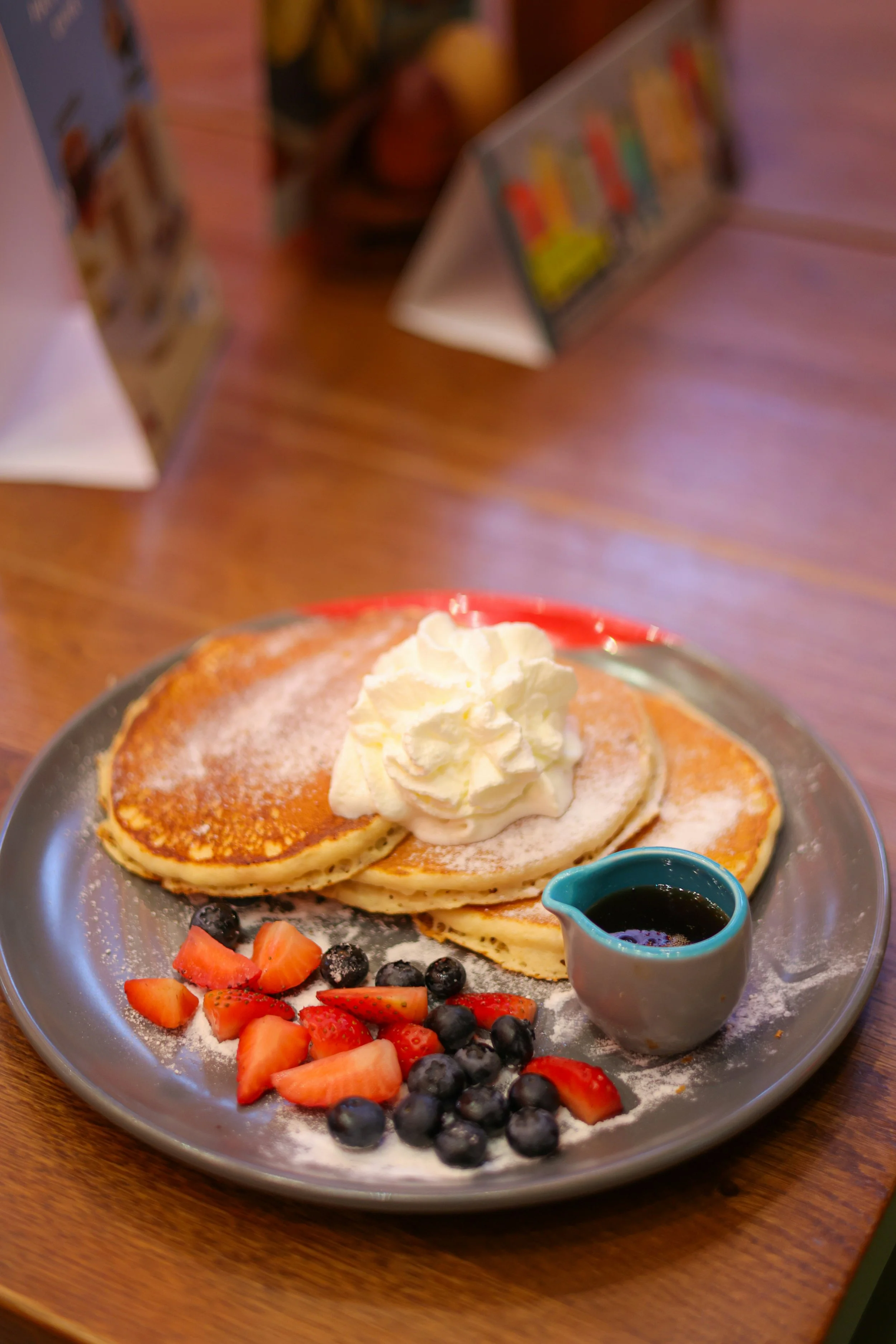 Plate of three pancakes topped with whipped cream, served with fresh strawberries and blueberries, a small pitcher of syrup, and dusted with powdered sugar on a wooden table.