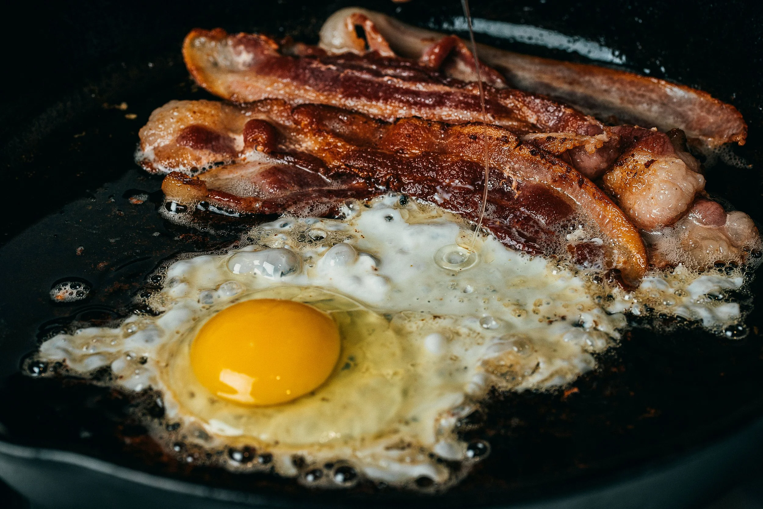 Bacon strips and a sunny side up egg frying in a skillet.