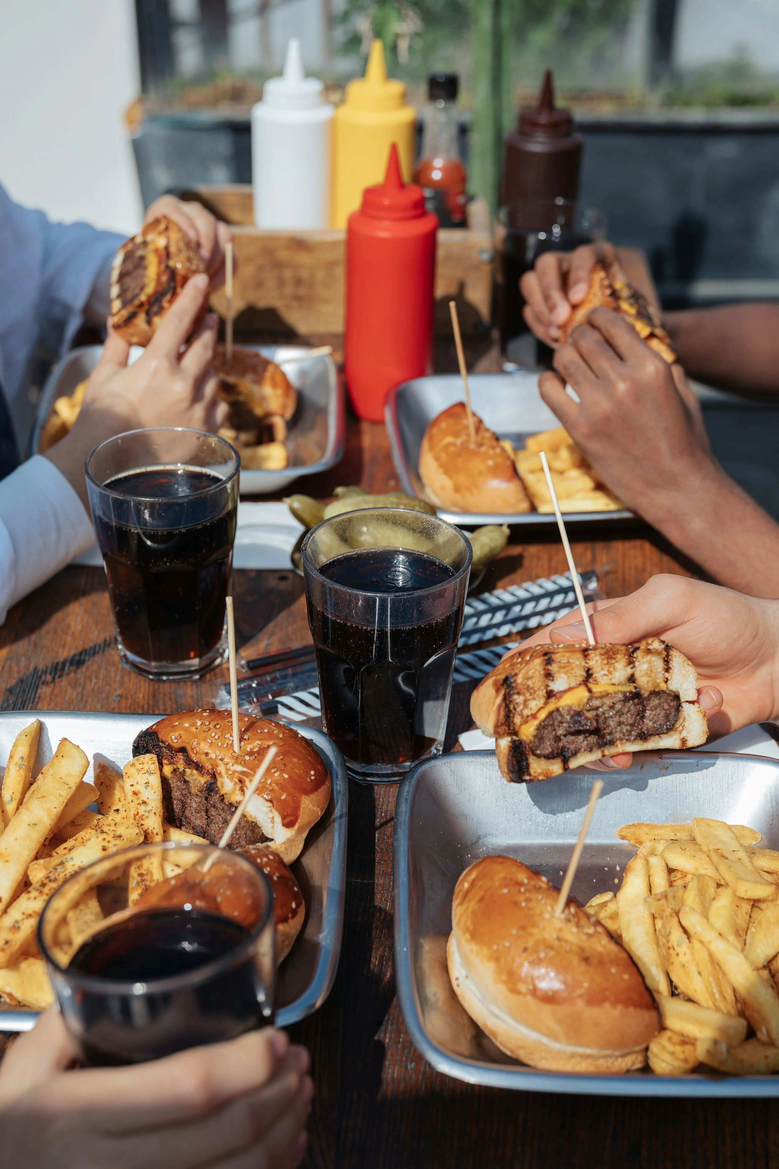 People enjoying burgers, fries, and soda at a wooden table with condiments in the background.