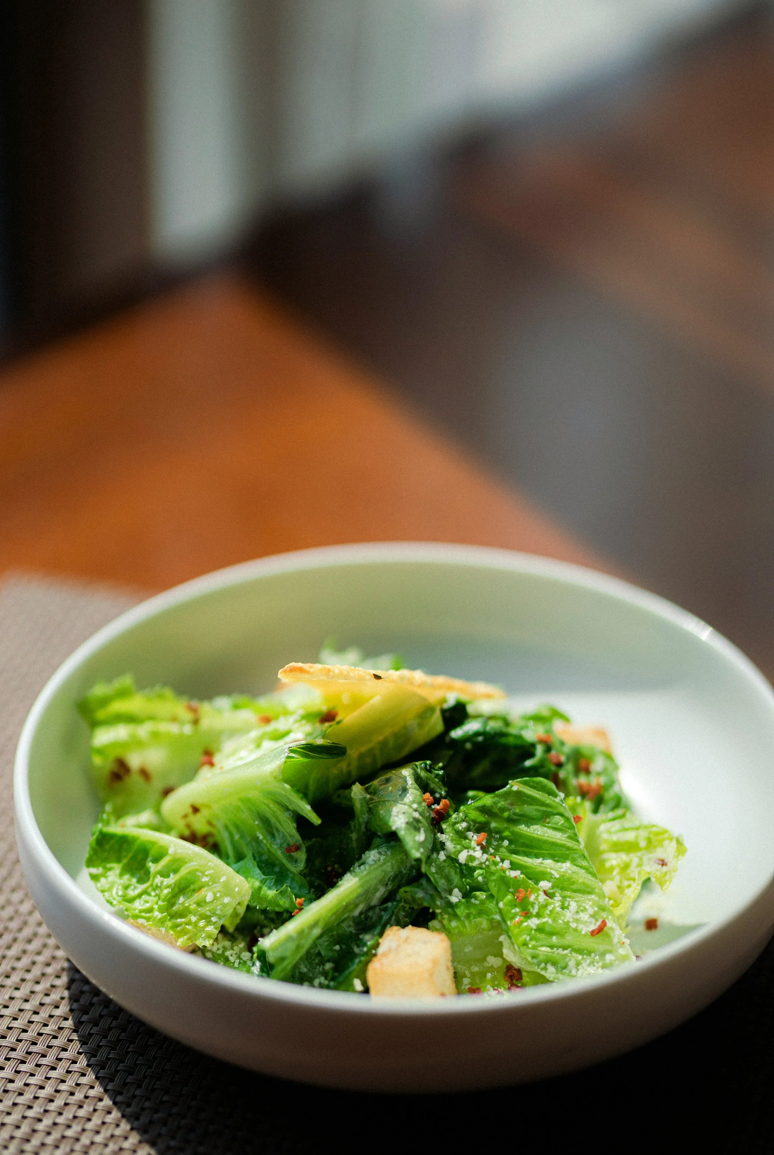 A bowl of Caesar salad with romaine lettuce, croutons, grated cheese, and black pepper on a textured placemat.