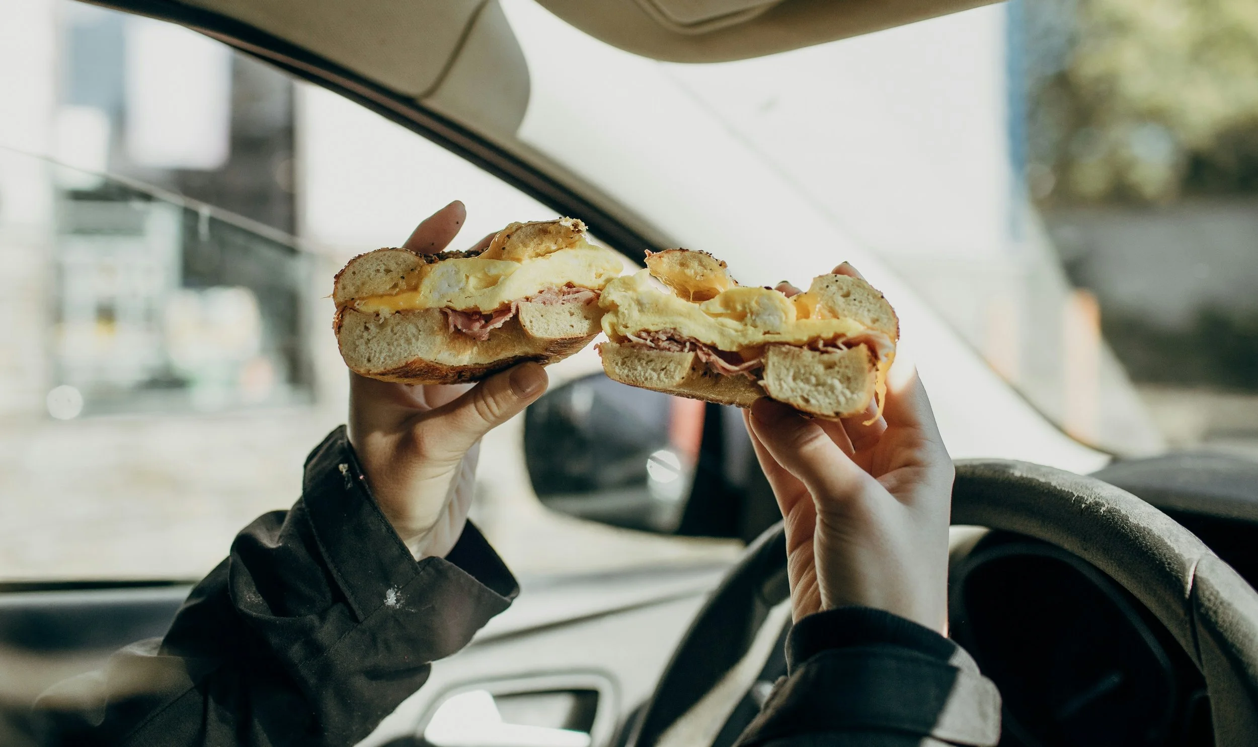 Person driving a vehicle holding two halves of a sandwich with melted cheese and meat inside.