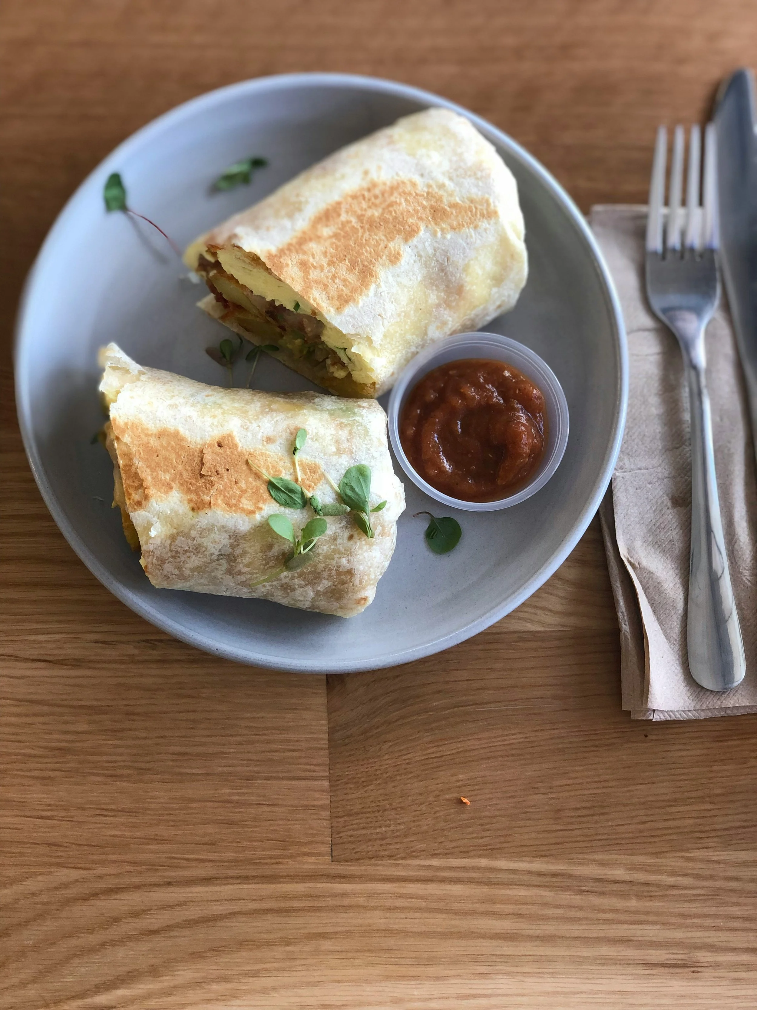 A plate with two burritos, garnished with microgreens, and a small container of salsa, placed on a wooden table with a fork and knife on a napkin to the right.