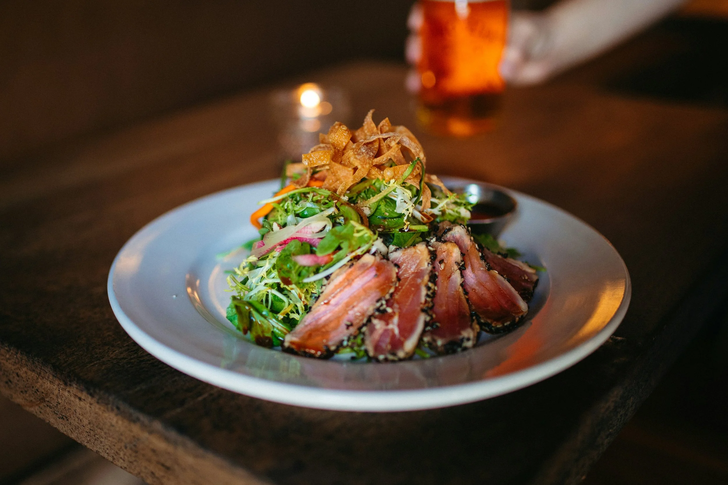 A white plate with slices of seared tuna coated in sesame seeds, topped with mixed greens and vegetables, and garnished with crispy fried wonton strips, served with a side of soy sauce, on a dark wooden table.