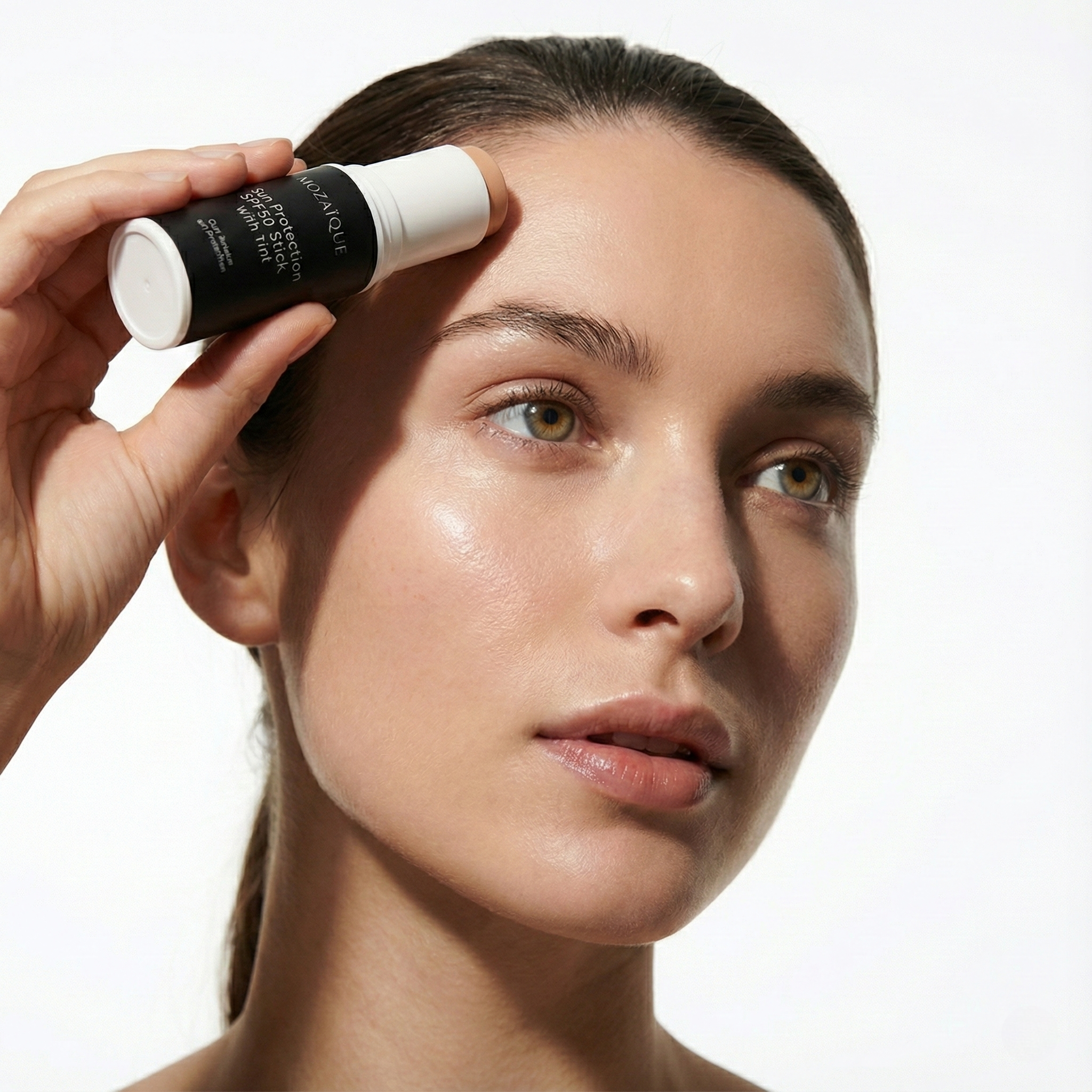 Close-up of a woman with fair skin and hazel eyes applying sunscreen to her forehead using a small bottle of sunscreen spray.