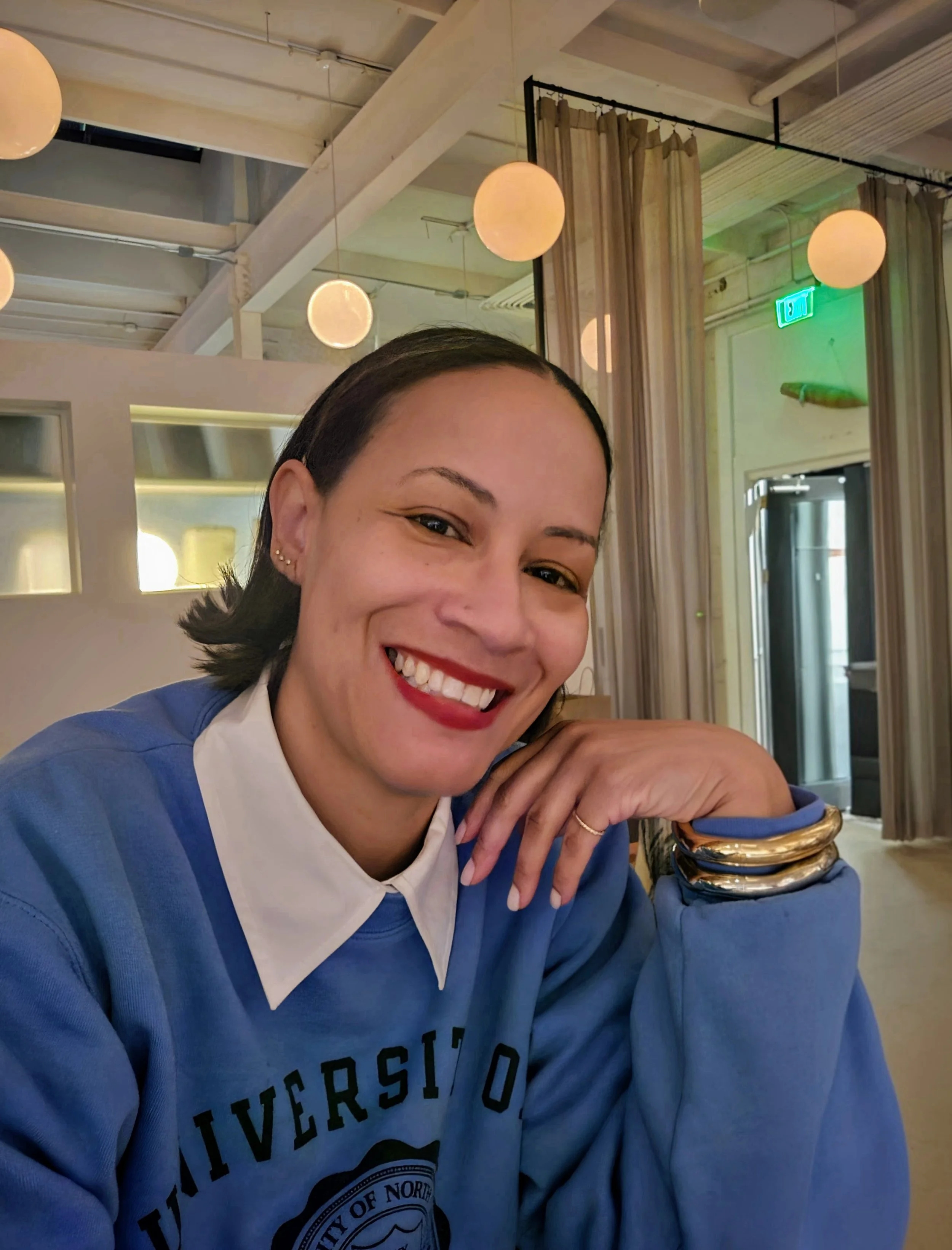 Smiling woman with dark hair, wearing a blue sweatshirt with text, leaning on her hand with gold and silver bangles, in a restaurant or cafe with hanging round lights and curtains.