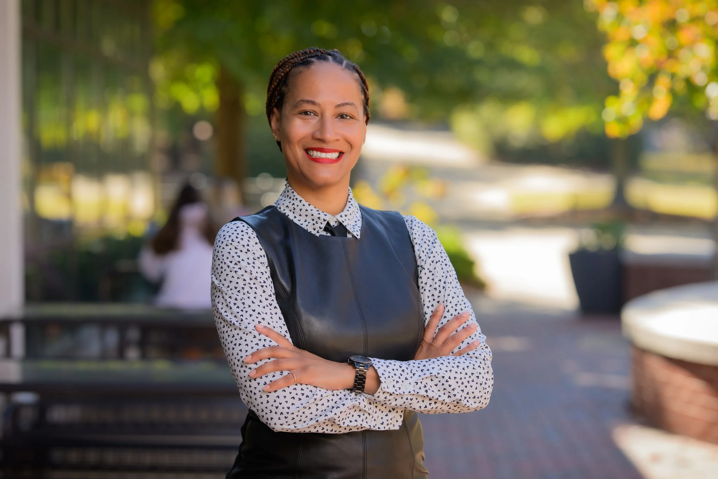 A woman with short braided hair smiling confidently outdoors in sunlight, wearing a patterned blouse and black leather vest, crossing her arms.