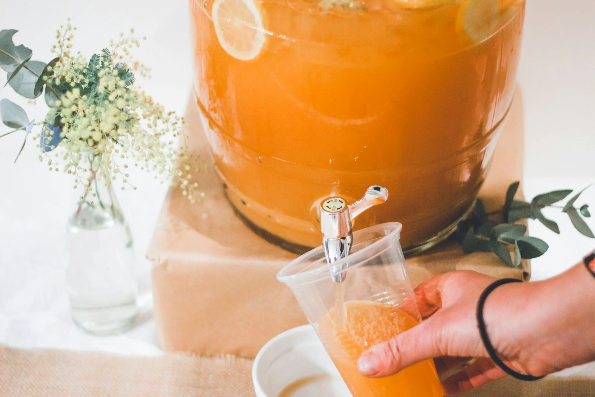 A clear plastic cup containing fruit-infused water with lemon, lime, strawberry, blueberry, and pineapple slices, with a straw, surrounded by fresh fruit slices and mint leaves.