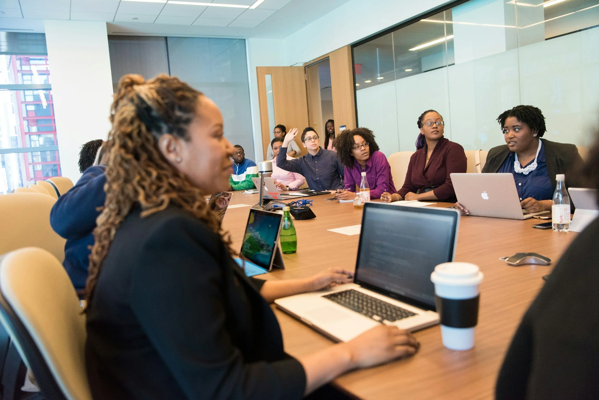 A diverse group of people seated around a conference table in a modern office, engaging in a meeting. Some are using laptops, and one person is raising their hand.