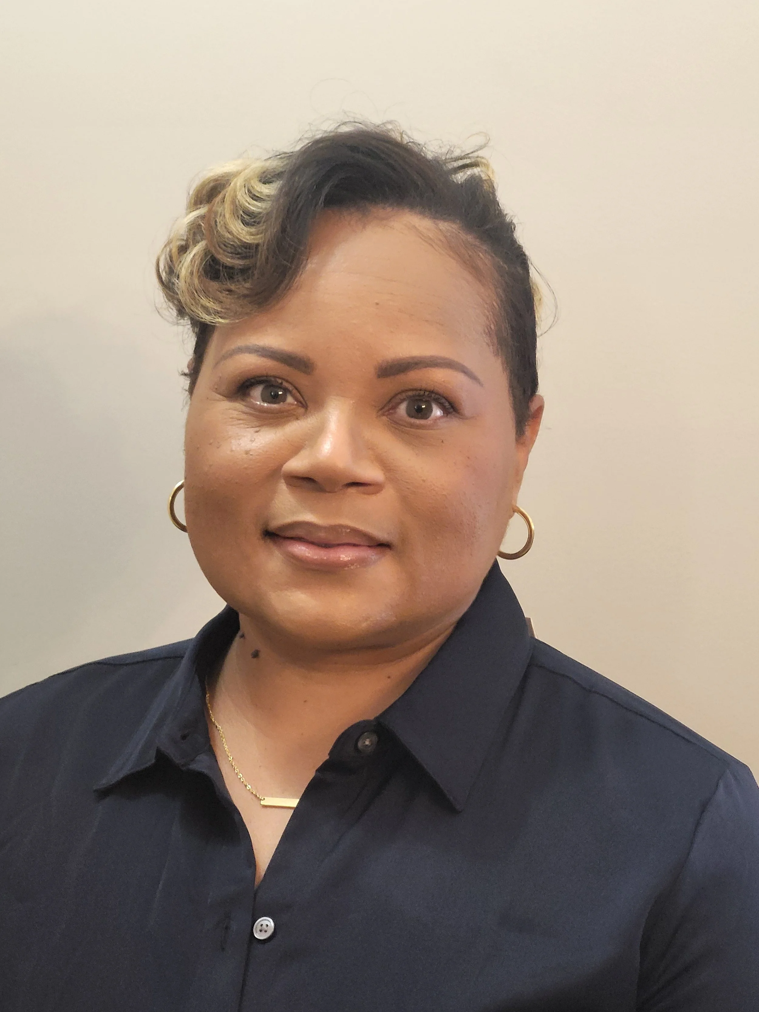 A woman with short curly hair styled with blonde highlights, wearing makeup, gold hoop earrings, a gold necklace, and a black collared shirt, standing against a beige wall.