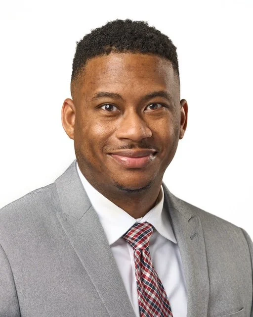 Headshot of a young African American man in a gray suit, white shirt, and red patterned tie, smiling against a white background.