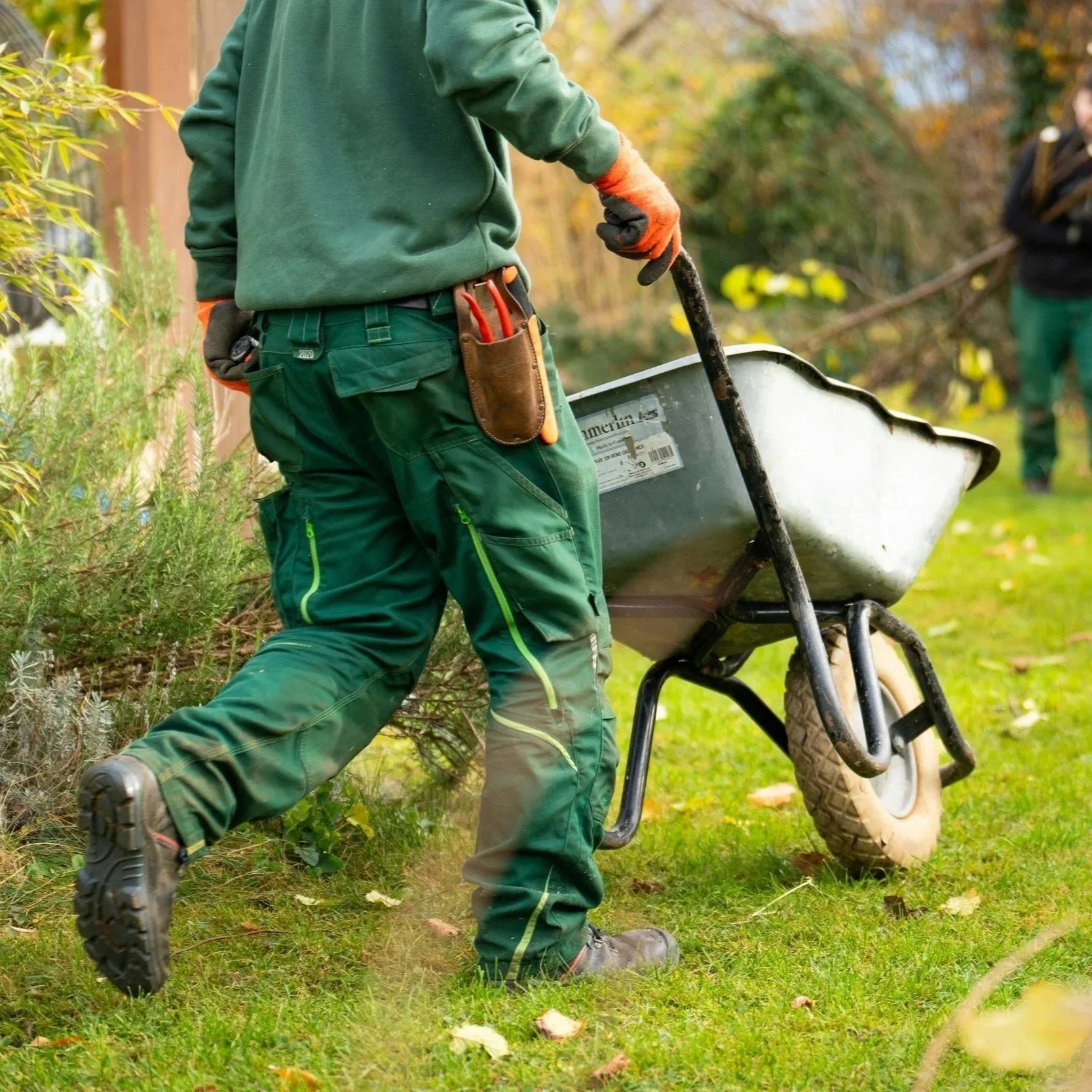Person wearing green outdoor clothes and gloves pushing a wheelbarrow in a garden.