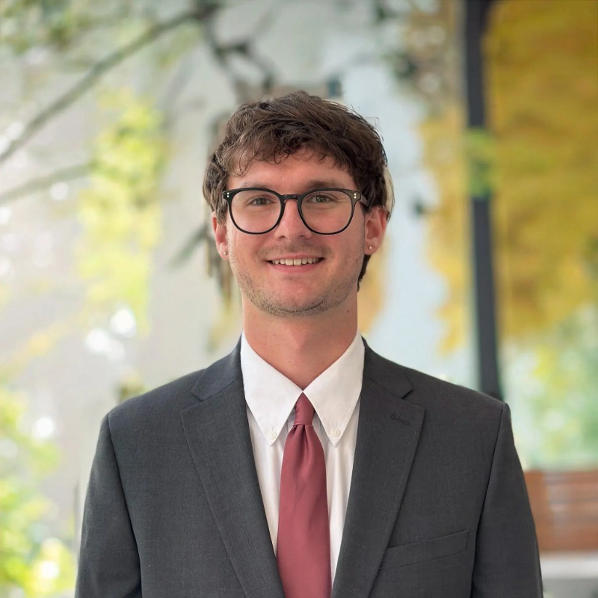 Headshot of a young man with brown hair, glasses, and earrings, wearing a dark suit, white shirt, and maroon tie, standing outdoors with blurred green and yellow foliage in the background.