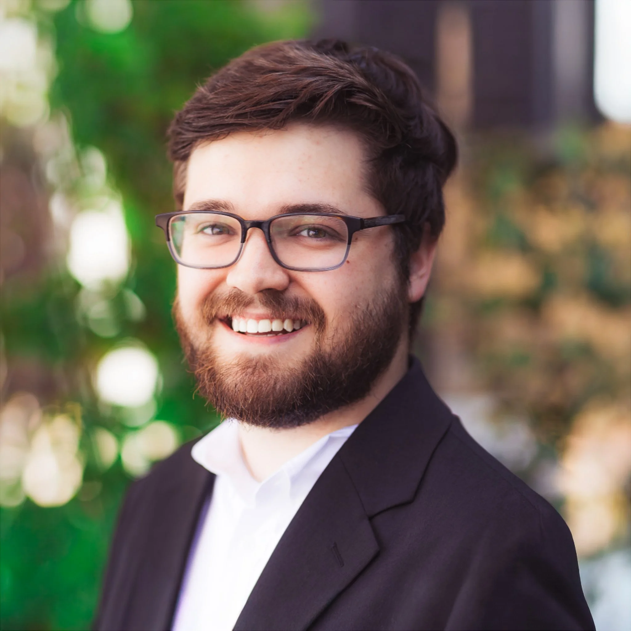 A smiling man with glasses and a beard, wearing a suit and white shirt, standing outdoors with a blurred green background.