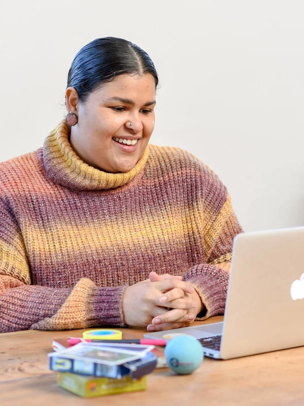 A woman with dark hair in a bun, wearing a multicolored turtleneck sweater, sitting at a wooden table with a silver MacBook, colorful small toys, and a blue spherical object, smiling and looking at her laptop.