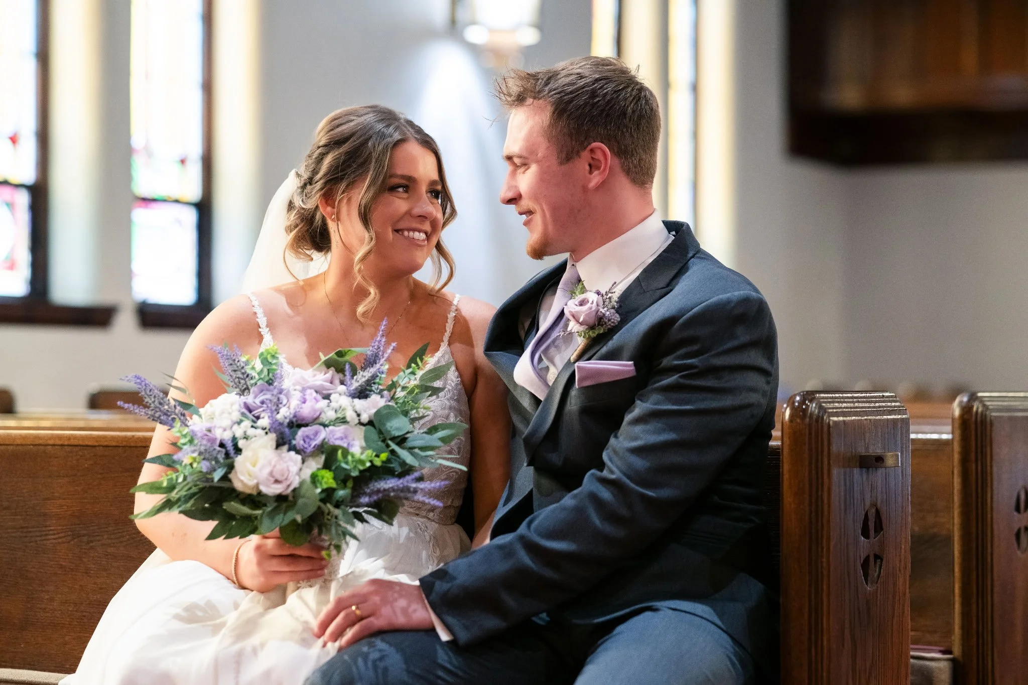 Bride with purple and white bouquet sitting next to groom in a church pew, looking into each other's eyes.ky.