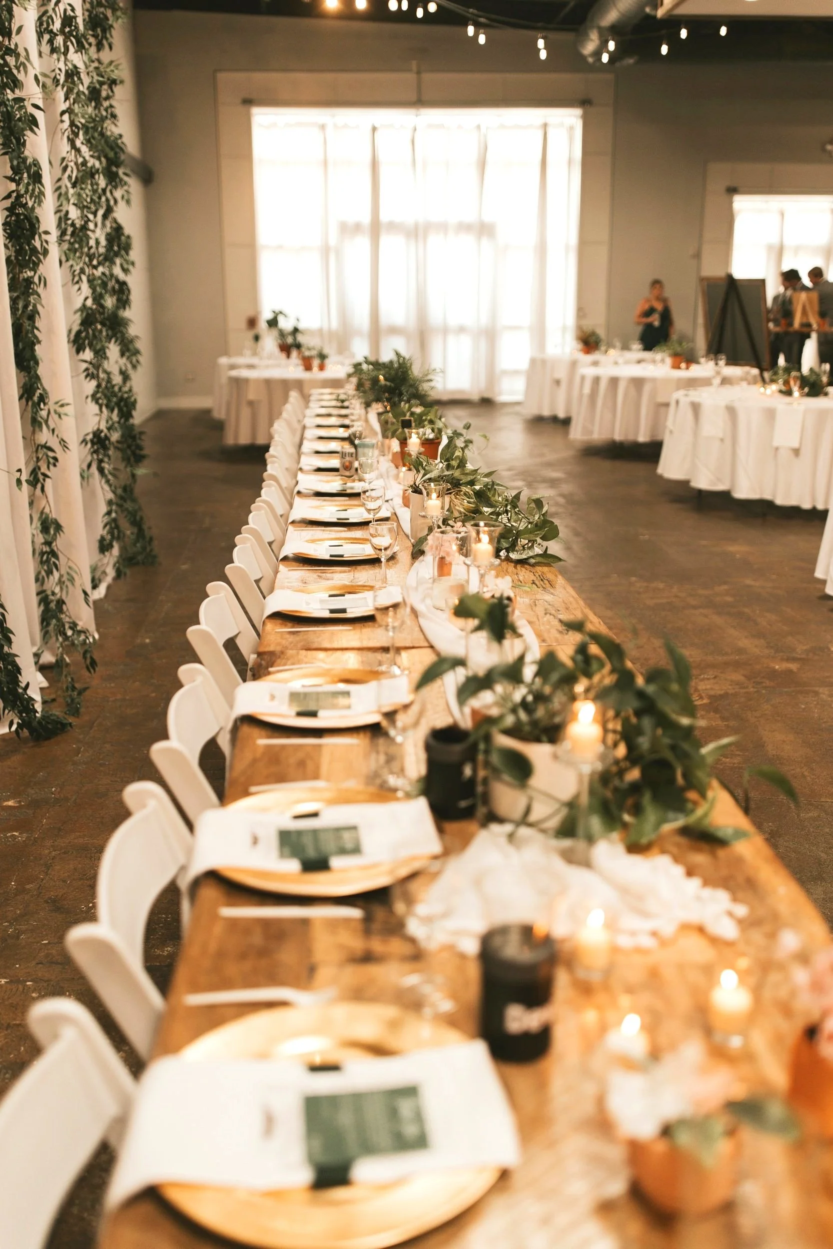 Long rectangular wooden banquet wedding head table set with gold charger plates, silverware, wine glasses, candles, and green foliage decor, in a brightly lit room with large windows and white walls. Minimalistic wedding table. Affordable, luxe.