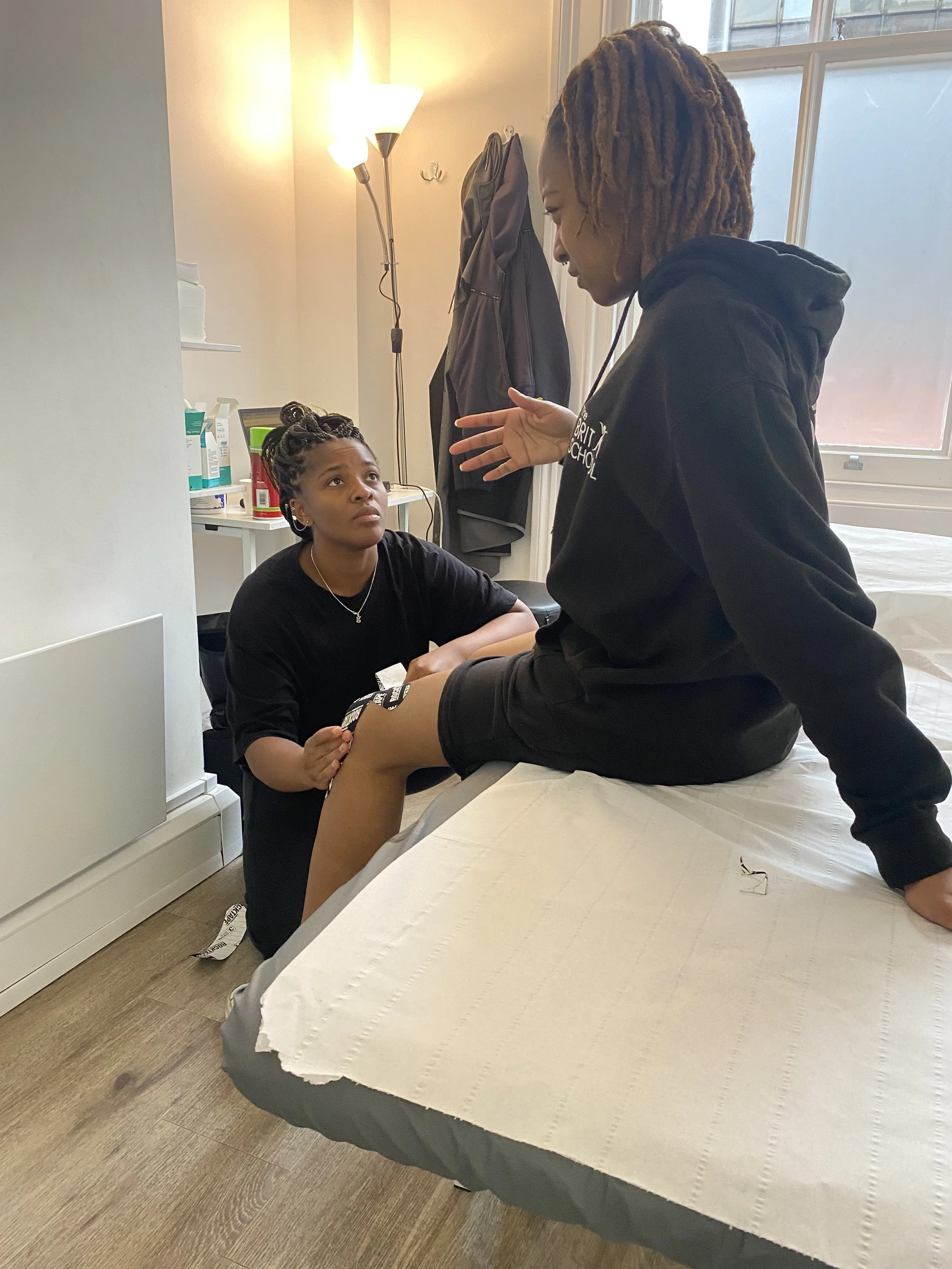 A healthcare worker or therapist is speaking with a patient who is sitting on a hospital bed, having a conversation indoors with medical supplies visible in the background.