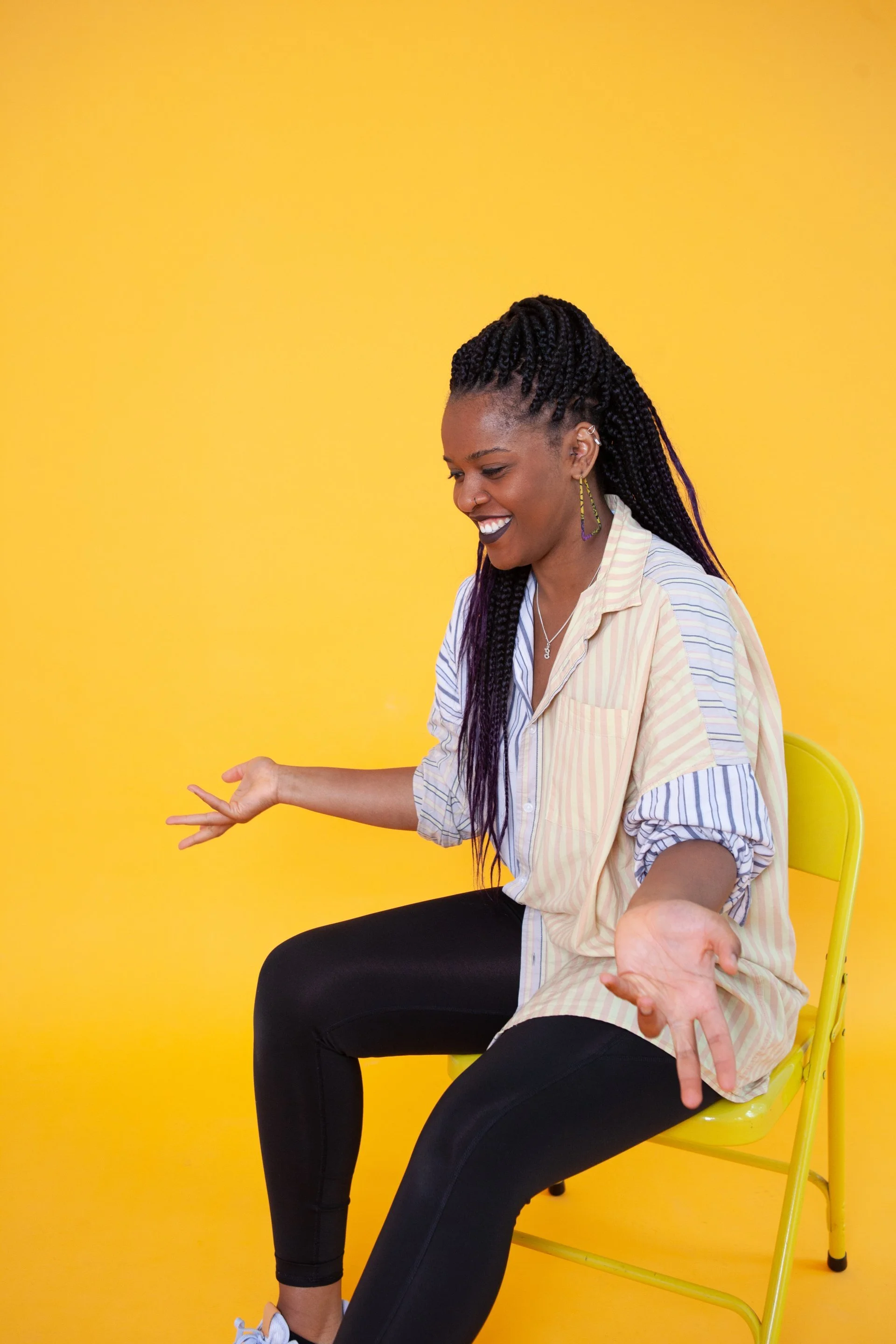 A woman with braided hair, wearing earrings and a striped shirt, sitting on a yellow chair against a yellow background, smiling and gesturing with her hands.