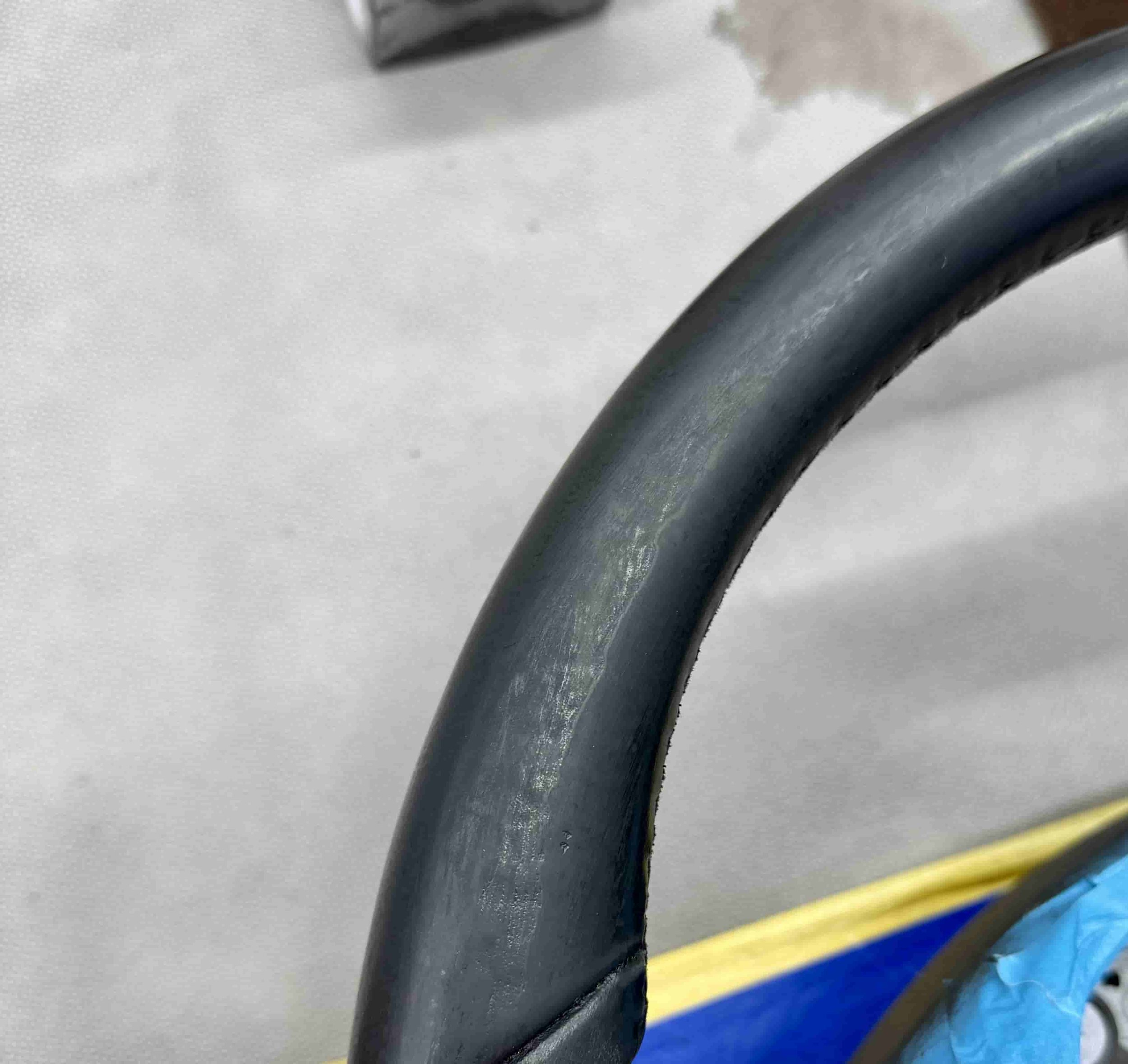 Nahaufnahme eines schwarzen Fahrradlenkers mit Gebrauchsspuren und grünlicher Ablagerung. Im Hintergrund sind Stoffe und Gegenstände erkennbar.