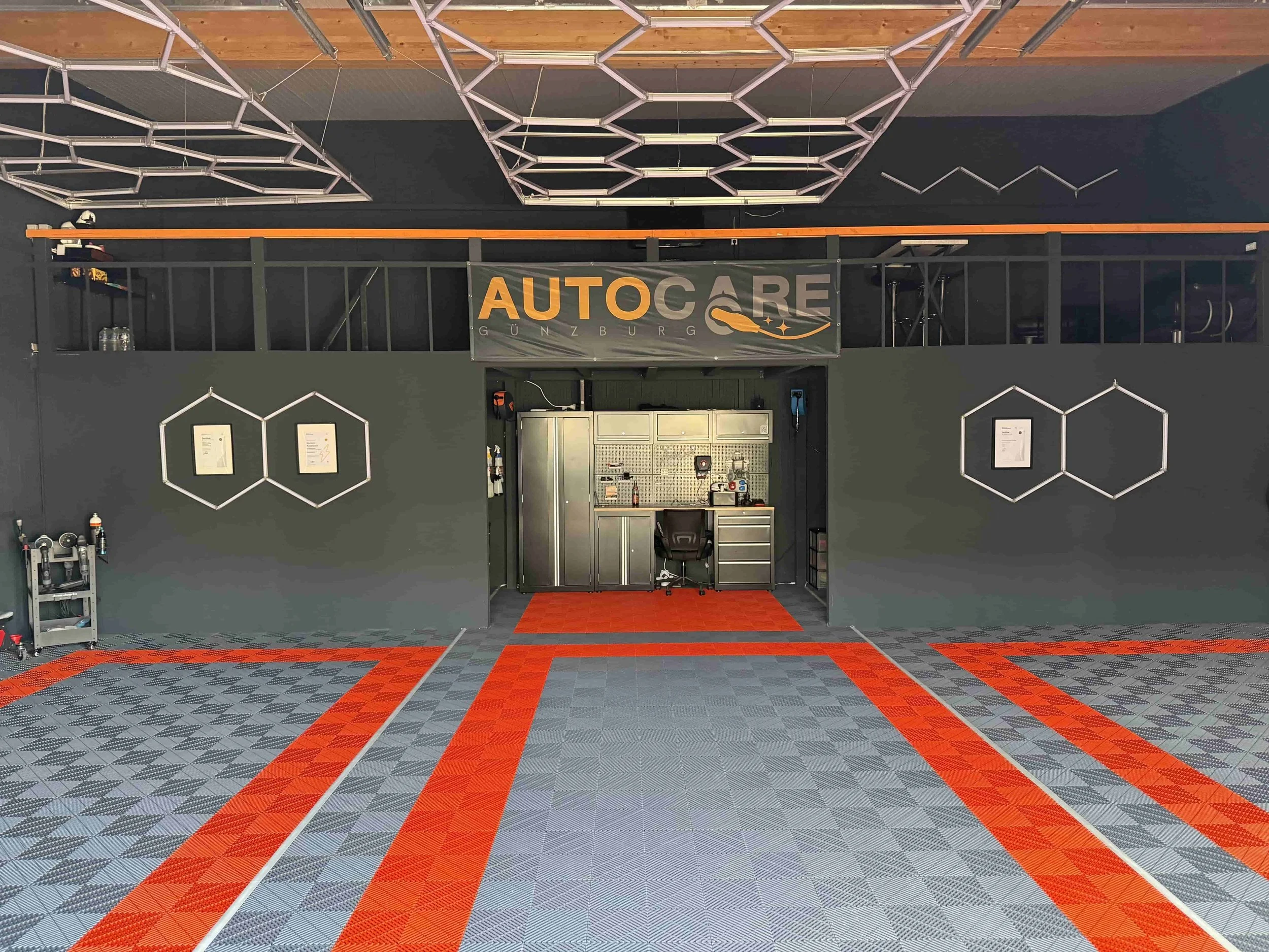 Ein modern eingerichteter Autowerkstattraum mit Werkstattelementen und einem ansprechenden, geometrischen Bodenmuster in Grau und Rot. Es gibt eine Arbeitsstation mit Werkzeugen, die auf einer Metallwand angebracht sind, und einen Firmenbanner mit dem Namen 'AUTOCARE Günzburg' hängt an der oberen Wand.
