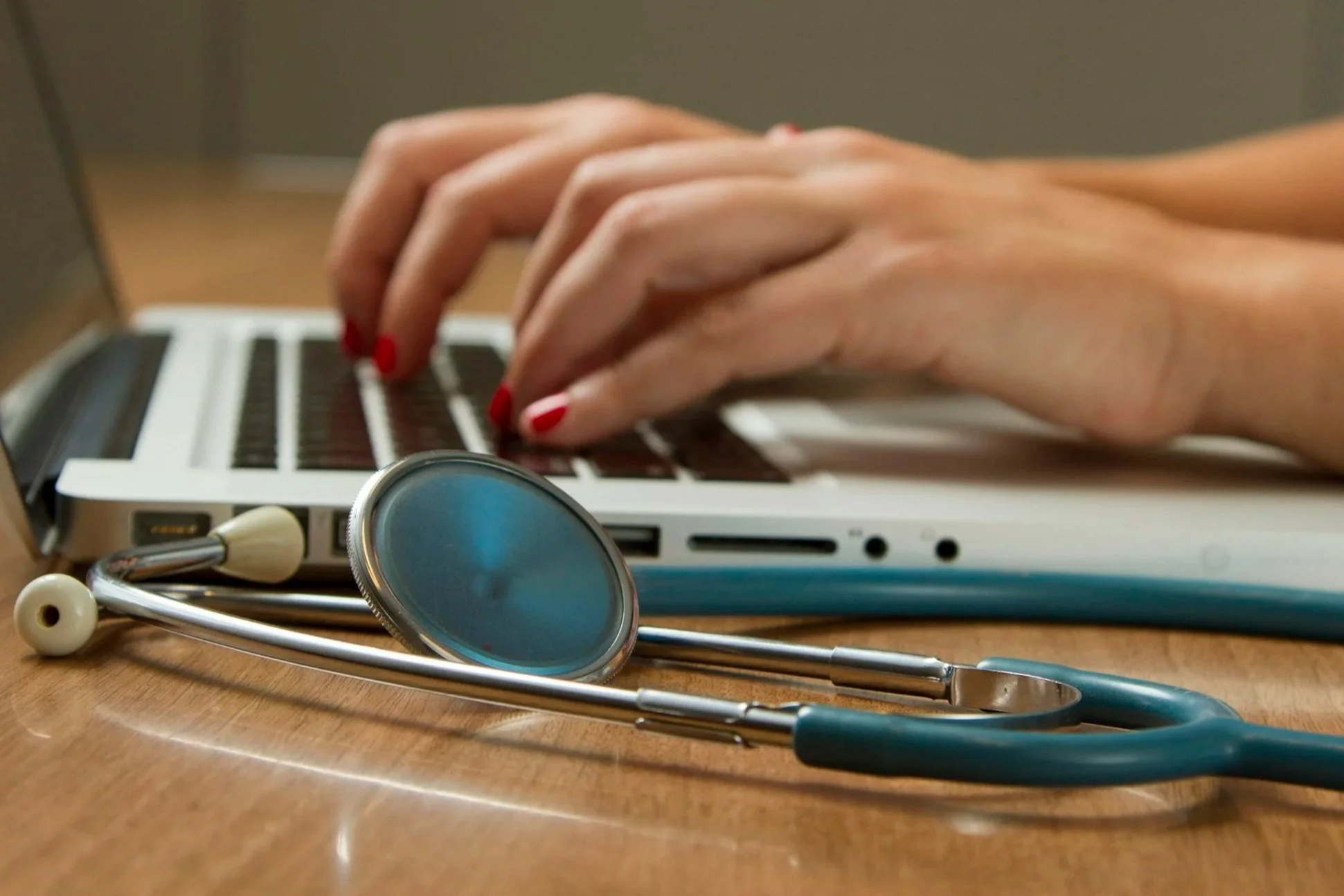 A stethoscope, a makeup brush, and a small mirror are placed on a wooden surface in front of a laptop keyboard. A person's hand with red nails is typing on the laptop.