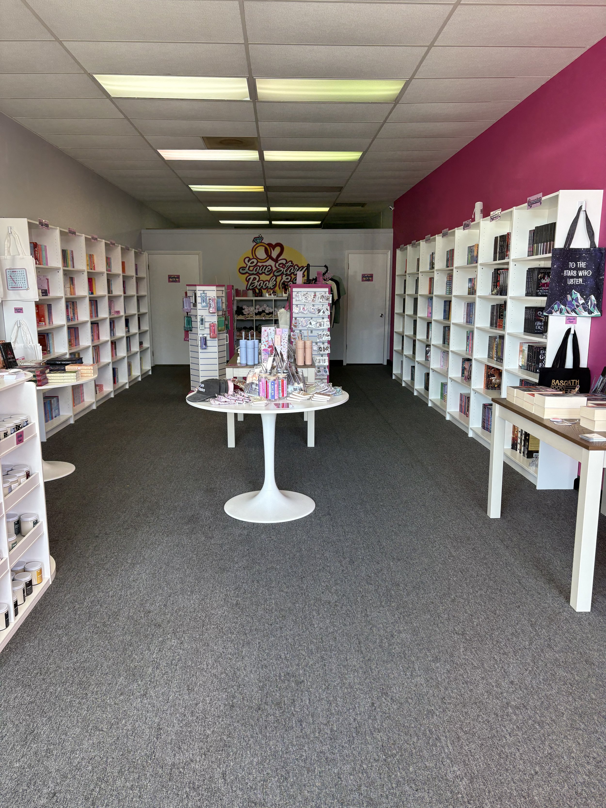 Photo of the store. On the side walls are shelves lined with books. In the middle are tables with book displays. The back wall has a shelving unit with t-shirts in the cubbies. Above the shelf is the store logo "Love Story Book Co.".