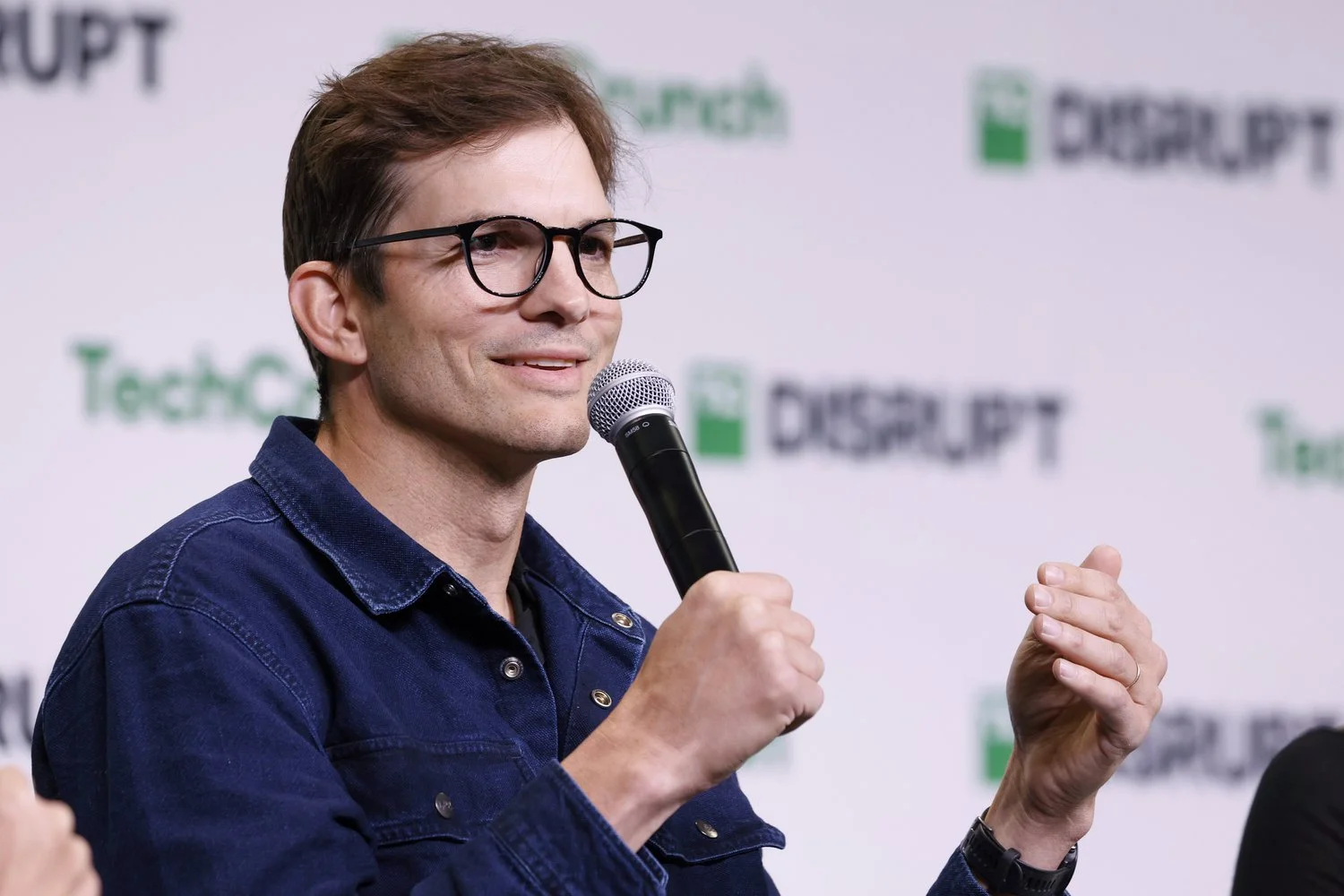 A man wearing glasses and a dark blue denim jacket speaking into a microphone at a TechCrunch event.