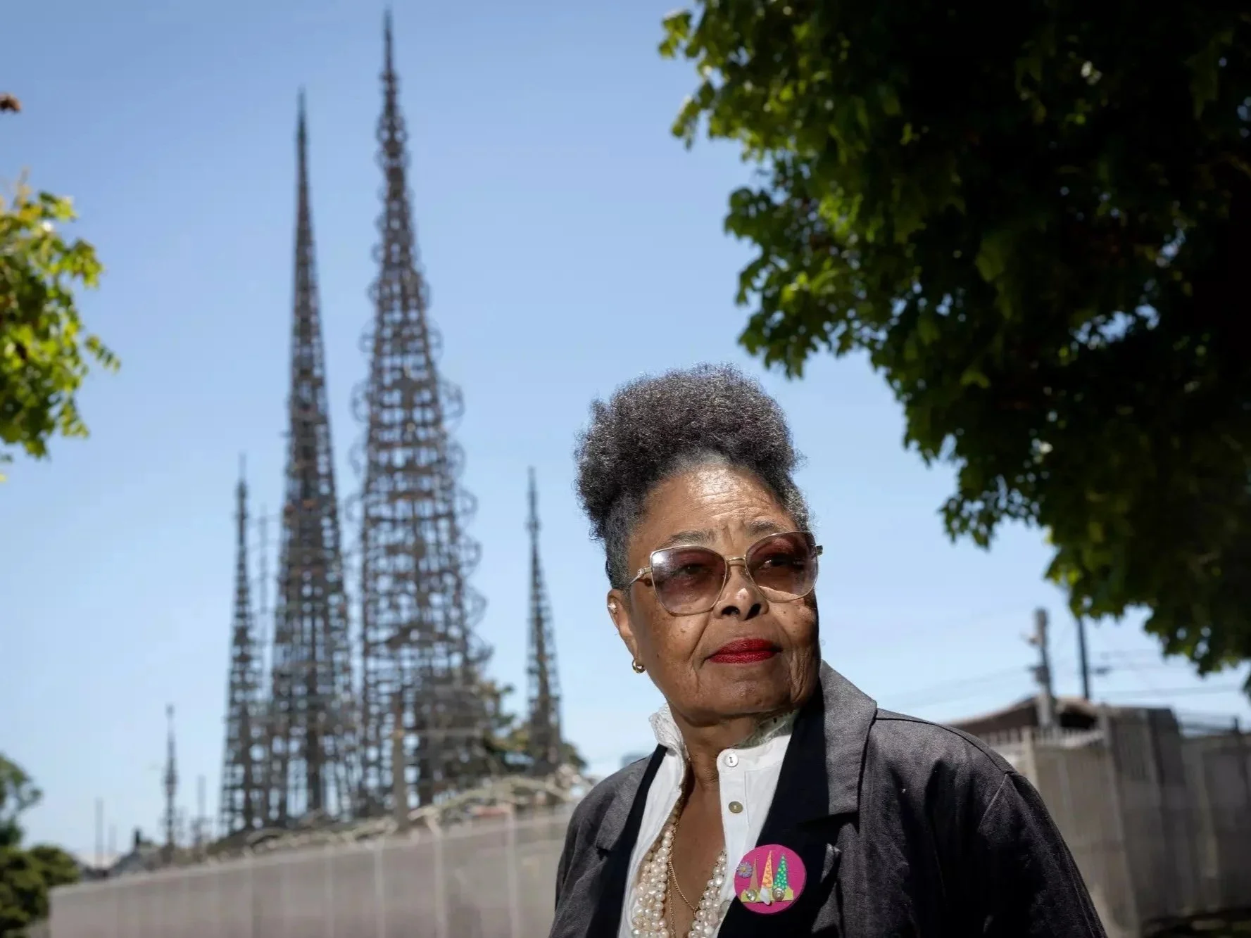 An elderly woman with glasses and short curly hair wears a pearl necklace and a gray blazer. She has a serious expression and wears a pink button with a colorful design on her blazer. Behind her are tall radio towers against a blue sky, with some green trees visible on the sides.