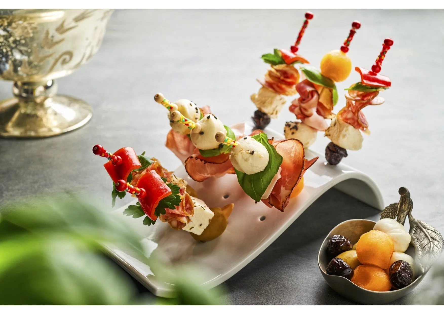 Skewers with various meats, cheese, and vegetables garnished with herbs and colorful toothpicks, presented on a white serving dish, with a bowl of fruit and a silver chalice in the background.
