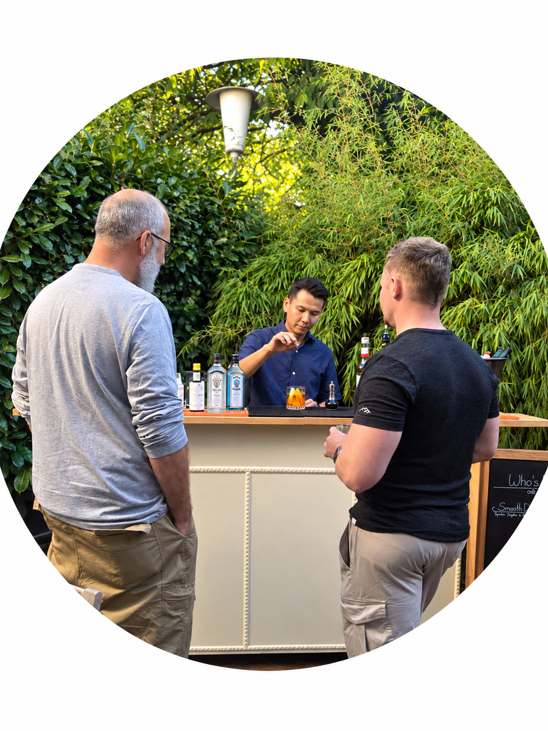 Three men at an outdoor bar with lush greenery background, bartender preparing a drink, bottles of alcohol on the bar, and chalkboard menu.