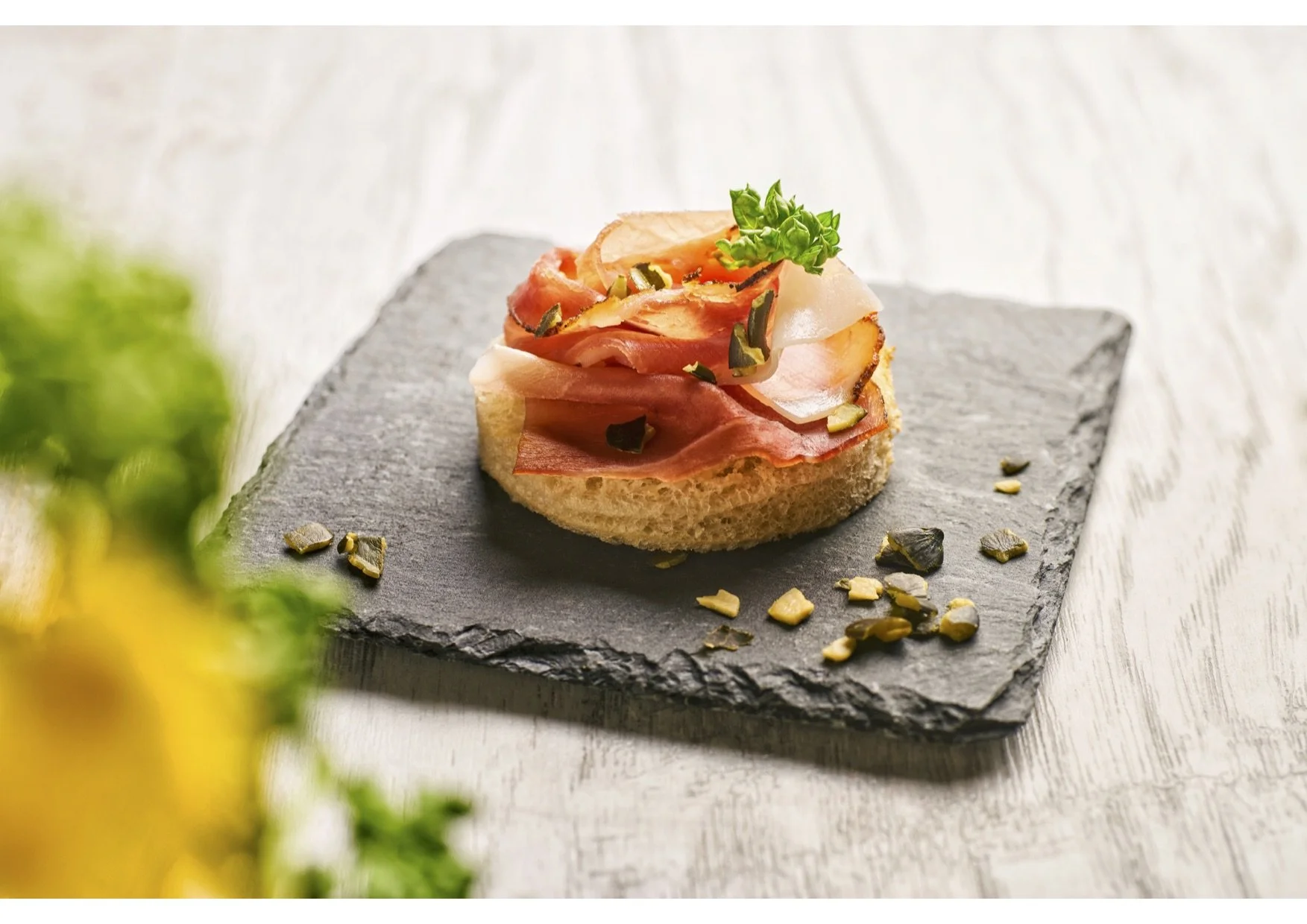An open-faced sandwich with ham, cheese, and herbs on a round piece of bread, garnished with parsley and sprinkled with seeds, served on a dark slate board.