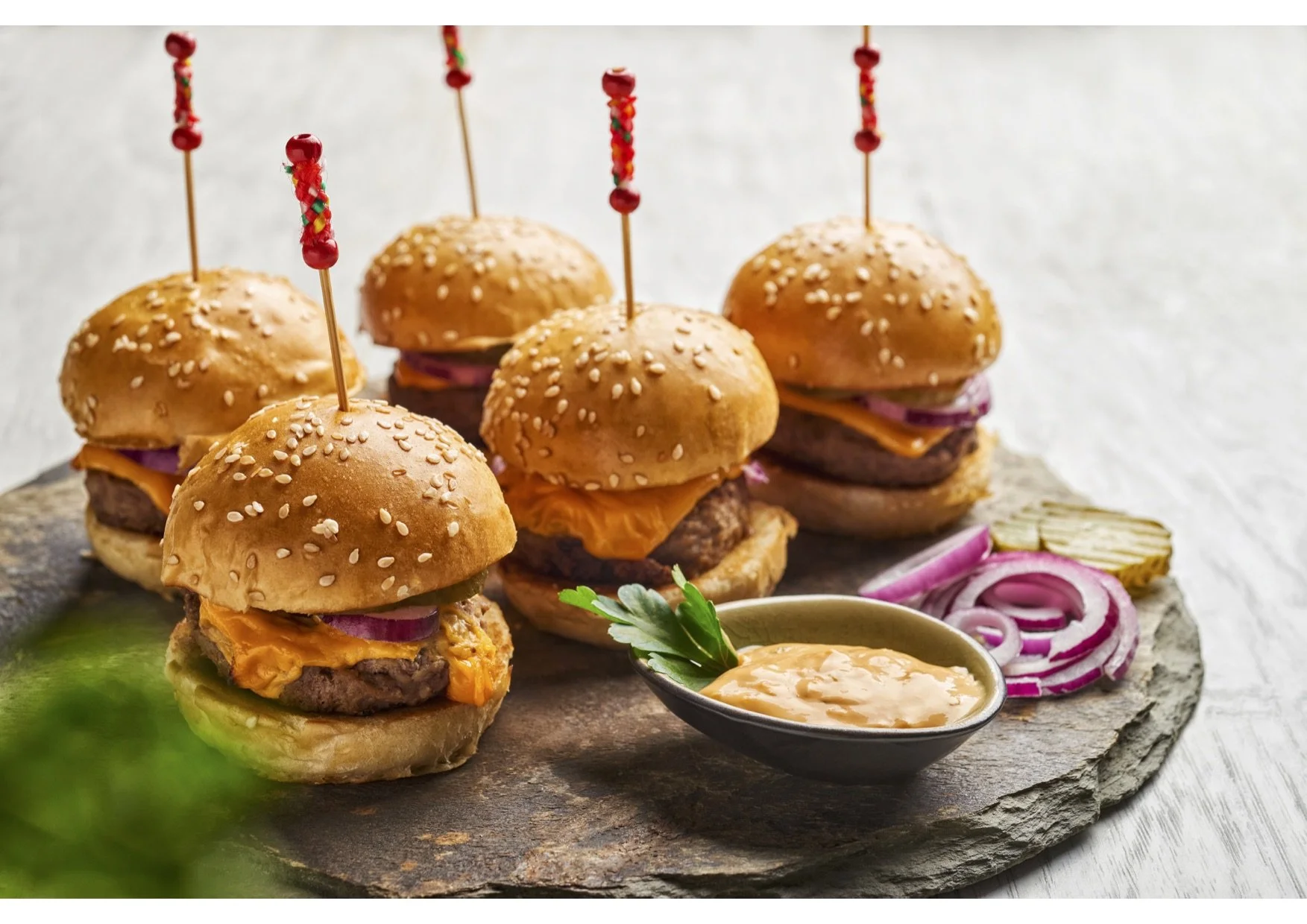 Four mini cheeseburgers with sesame seed buns, lettuce, onion, cheese, and beef patties, garnished with parsley, served with sliced onions and a creamy dipping sauce on a round stone platter.