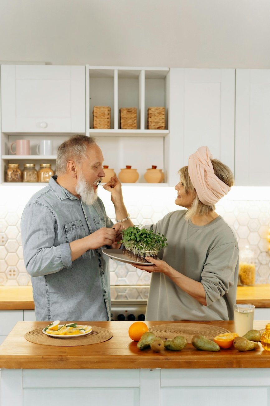 A woman giving a piece of food to a man in a kitchen while holding a plate of fresh greens. The kitchen features white cabinets, jars, and decorative items on the shelves, with a wooden counter displaying fruits and drinks.