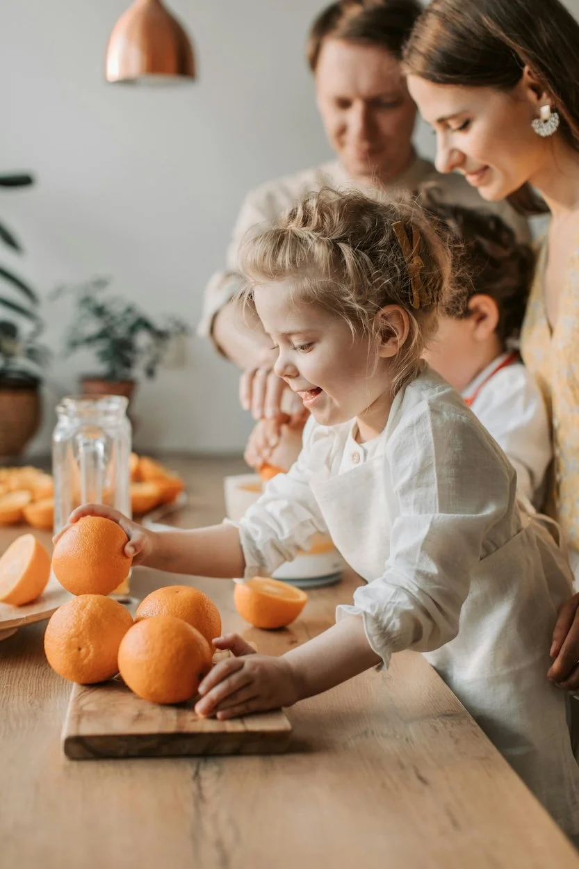 A young girl with curly hair is smiling while holding a halved orange, surrounded by whole oranges on a wooden table. Two adults and another child are in the background, all gathered in a bright kitchen or dining area.to represent who Bayside